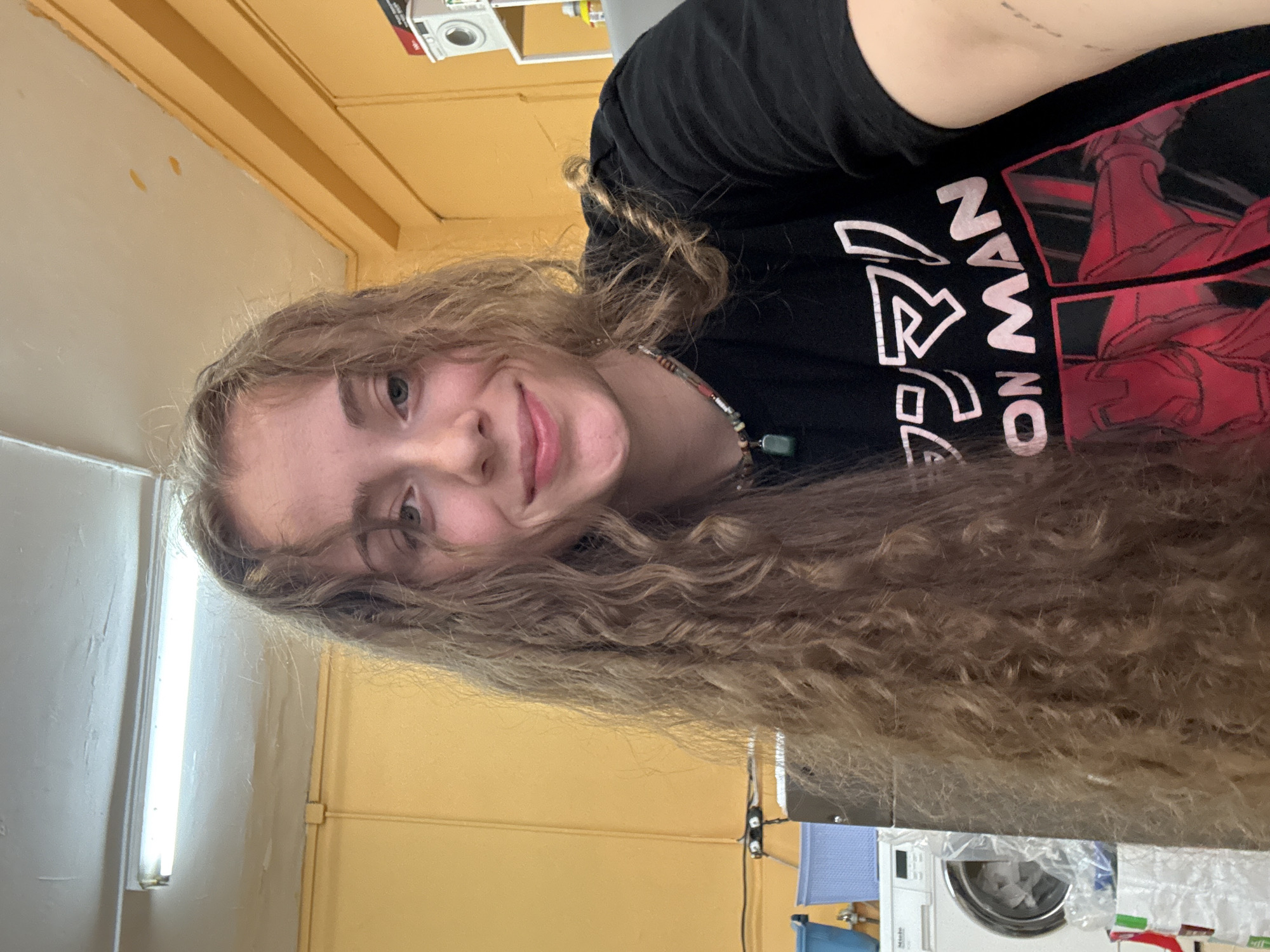 A young woman with long, curly brown hair smiles at the camera. She is wearing a black t-shirt with a graphic of Iron Man and Japanese text. She is also wearing a beaded necklace with a green stone pendant. The background appears to be an interior space with yellow walls and a fluorescent light fixture on the ceiling. A washing machine and laundry baskets are visible to the left of the woman. The location is the laundromat of a hostel in Sofia, Bulgaria.