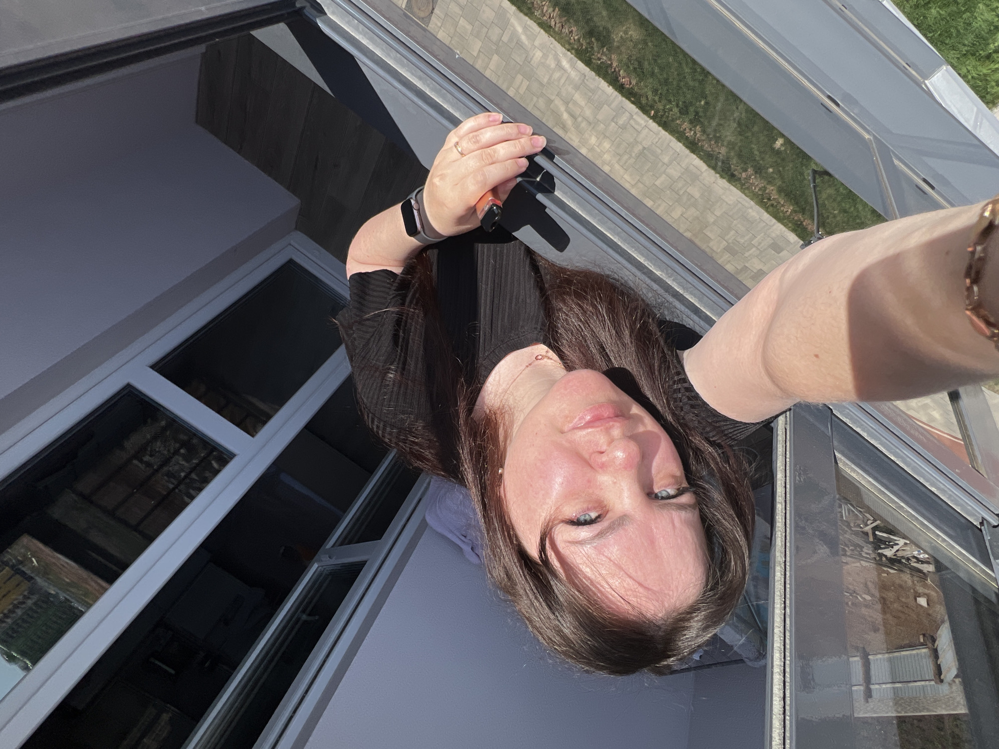 A woman with brown hair and blue eyes is taking a selfie while standing near a window. She is wearing a black shirt and a smartwatch. Her right arm is extended with the camera and her left hand is holding onto the window frame. The window frames are white. Through the windows, it is possible to see the reflection of the inside of a house, a brick building and a green lawn. The image appears to be taken outdoors on a sunny day. The specific location within Pervomaysky, Russia, is not discernible from the provided image.