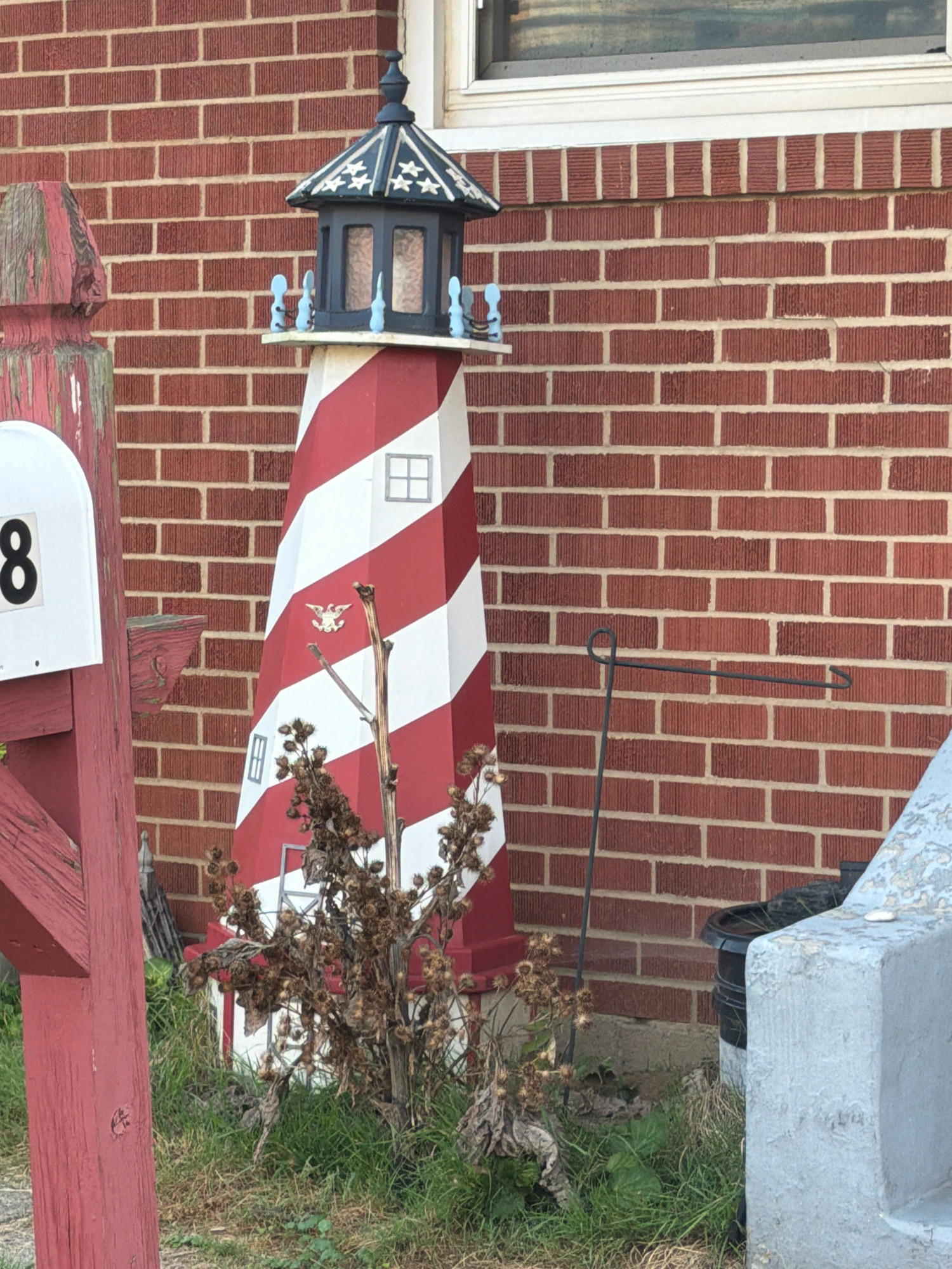 In Harmar Township, United States of America, a decorative lighthouse sits near a red brick wall. The lighthouse features red and white stripes and a dark blue top with stars. A white mailbox with the number "8" is visible to the left. A dry, thorny bush is in front of the lighthouse. A black trash can is partially obscured behind the lighthouse. A grey concrete structure is to the right of the scene. The ground is covered with grass and some dead leaves.