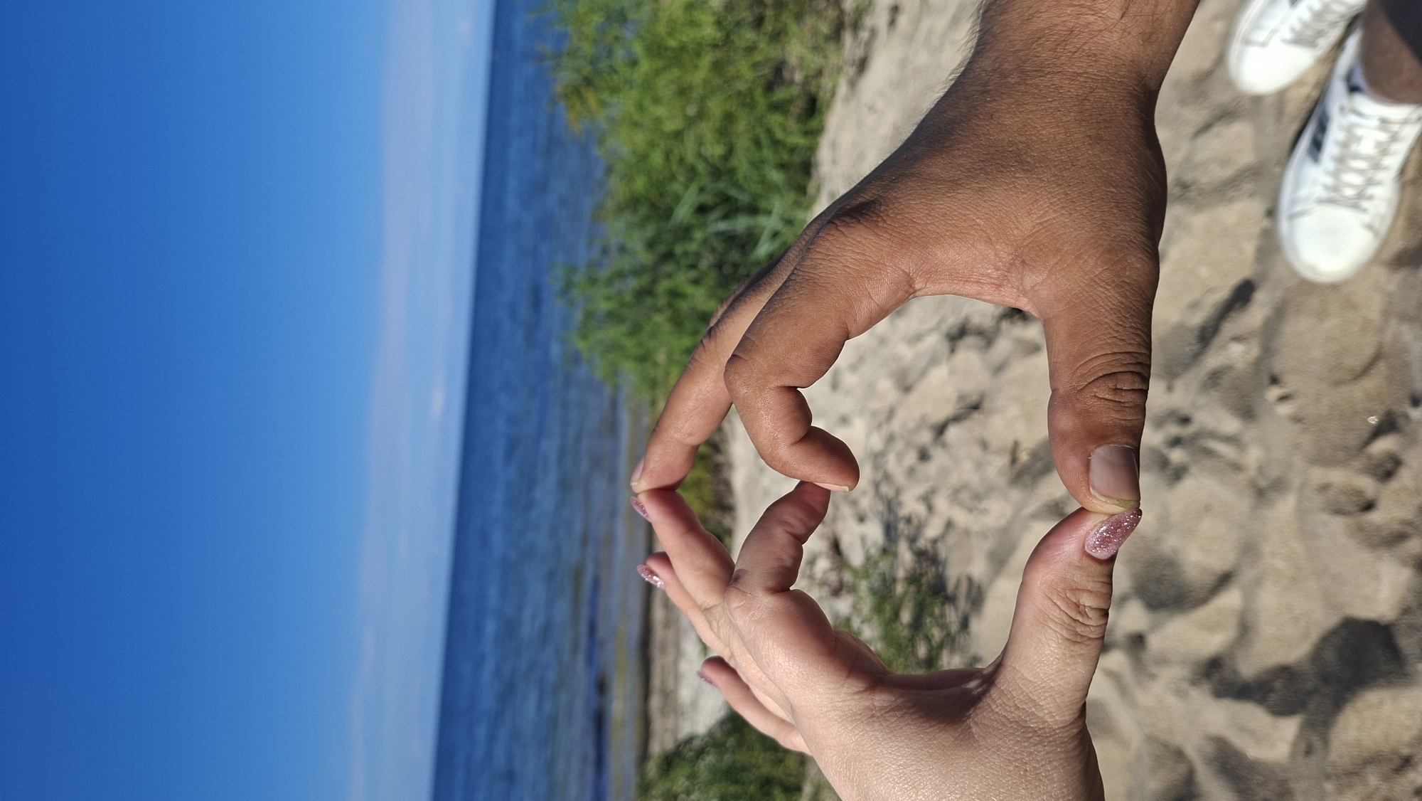 The image captures a close-up view of two hands forming a heart shape against a backdrop of the sea and a sandy beach, located in Meremõisa, Estonia. The left hand, belonging to a person with lighter skin, displays fingernails with glittery pink polish. The right hand, with darker skin, is positioned to complete the heart shape. The hands are the primary focus, positioned in the lower center of the frame. Behind the hands, a sandy beach stretches towards the water, with sparse green vegetation visible along the shoreline. The sea appears calm and blue, meeting a clear blue sky. In the lower right corner of the image, a portion of white sneakers is visible, suggesting the presence of at least one person. The overall scene evokes a sense of peace and connection.