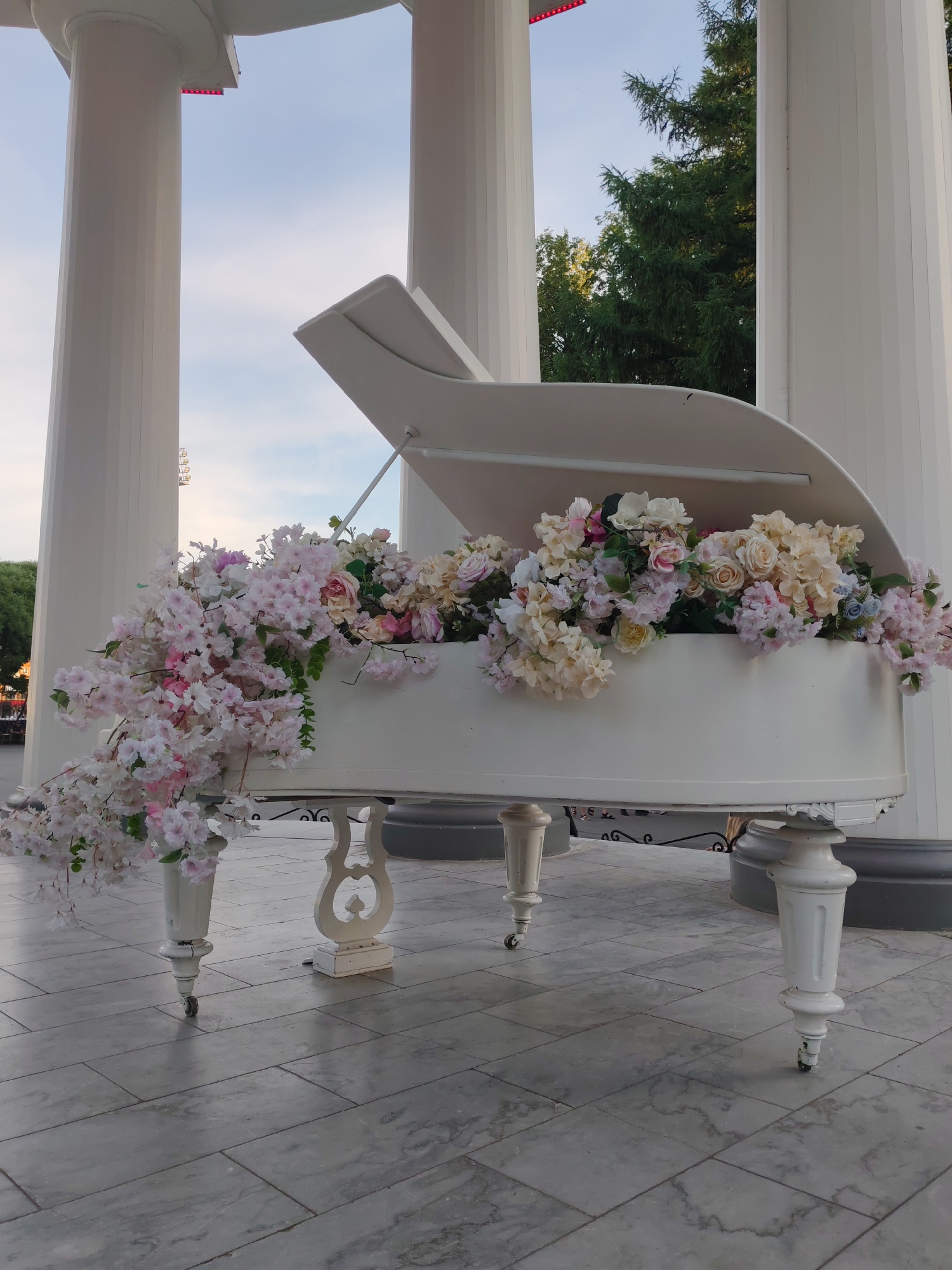 A white grand piano, adorned with an abundance of light pink and cream-colored artificial flowers, is positioned outdoors on a tiled patio. The piano is under a large white structure supported by thick, fluted columns. In the background, trees are visible against a pale blue sky. The scene appears to be a public space, possibly a park or the grounds of a venue. There are no people clearly visible in the image. The piano is decorated as if for a special event or as a permanent artistic installation. The overall impression is one of elaborate decoration and a serene, possibly celebratory, atmosphere. The location is the city of Perm, Russia.