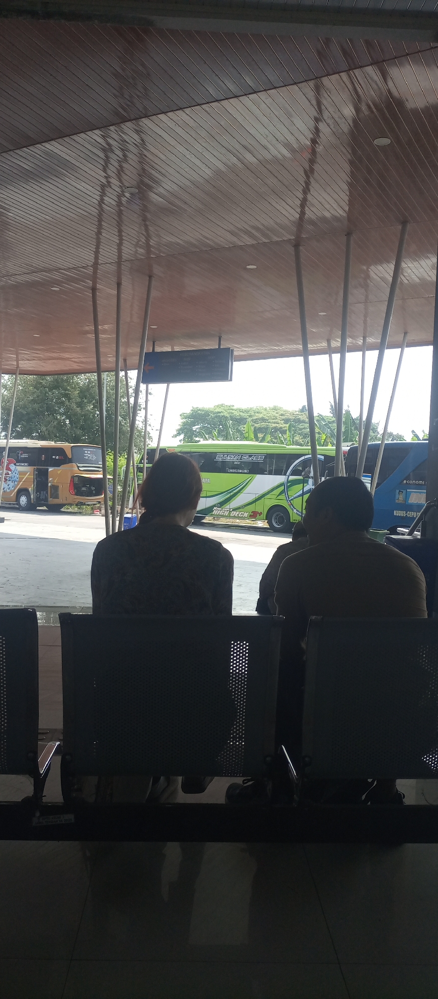 The image captures a scene at a bus terminal in Tambakromo, Indonesia. In the foreground, two individuals are seated on dark, perforated chairs, facing away from the camera. The person on the left has dark hair and wears a patterned shirt. The person on the right has short dark hair and wears a light-colored shirt. They appear to be waiting.
Behind them, the bus terminal is visible. A sign with text and an arrow is mounted on a pole above the waiting area. Two buses are parked outside. The bus on the left is yellow with a graphic design. The bus on the right is green and white with "SULTAN CLASS" and "HIGH DECK" written on its side. The terminal has a modern, open-air design with a sloped, wooden ceiling supported by multiple thin poles. Trees and foliage are visible in the background, suggesting an outdoor setting. The floor in the foreground is a dark, reflective surface, likely polished concrete or tile, reflecting the overhead structure and the light.
