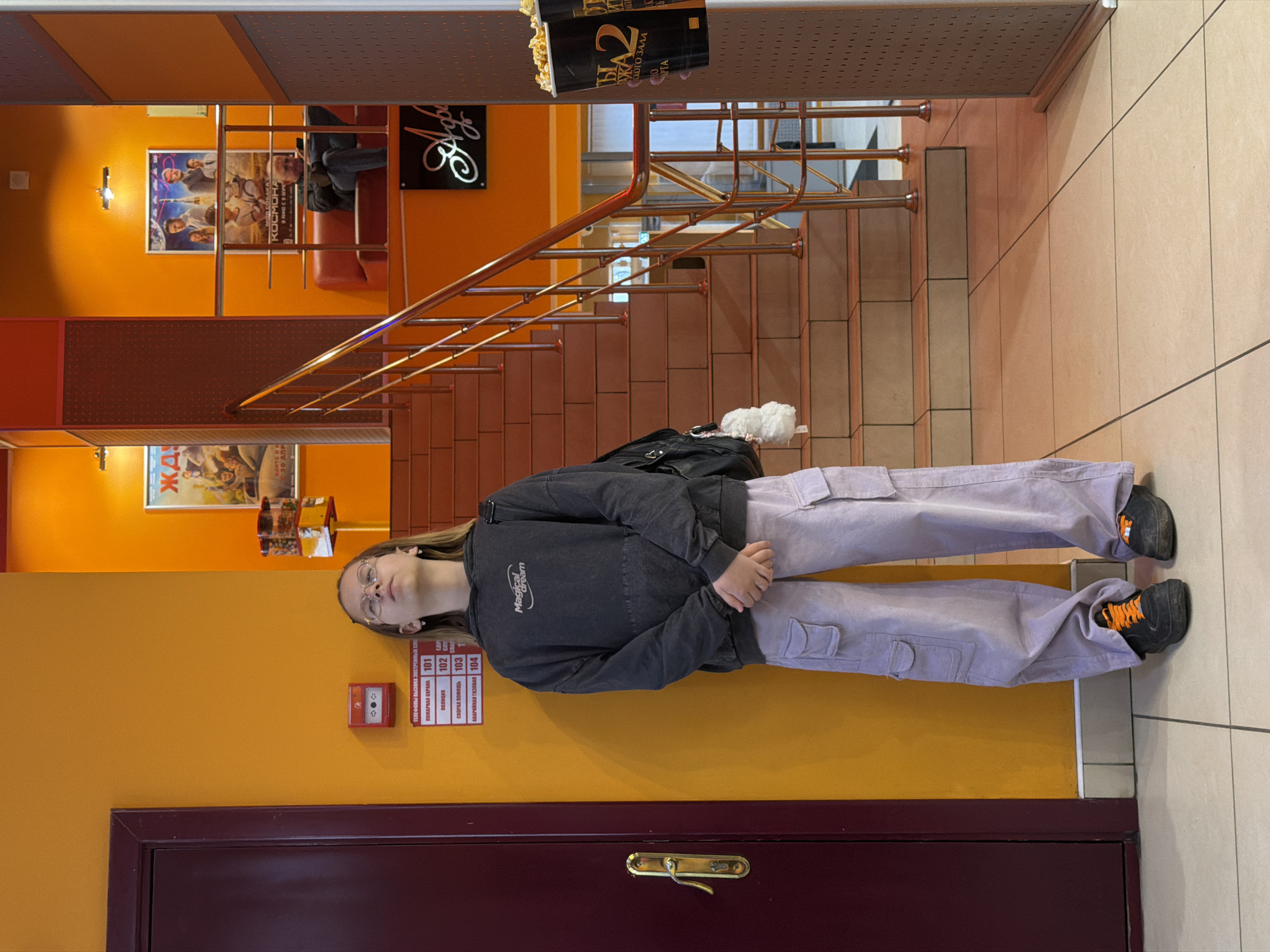 The image shows a young woman in light purple cargo pants and a dark jacket standing inside what appears to be a movie theater. She is wearing glasses and black shoes with orange details. The background features orange walls adorned with movie posters and safety information signs in Russian. A staircase with metal railings leads to an upper level with seating and decorative elements. A bucket of popcorn is visible near the top of the stairs. The floor is tiled in light colors. The scene is indoors, likely in the city of Unknown, Unknown.
