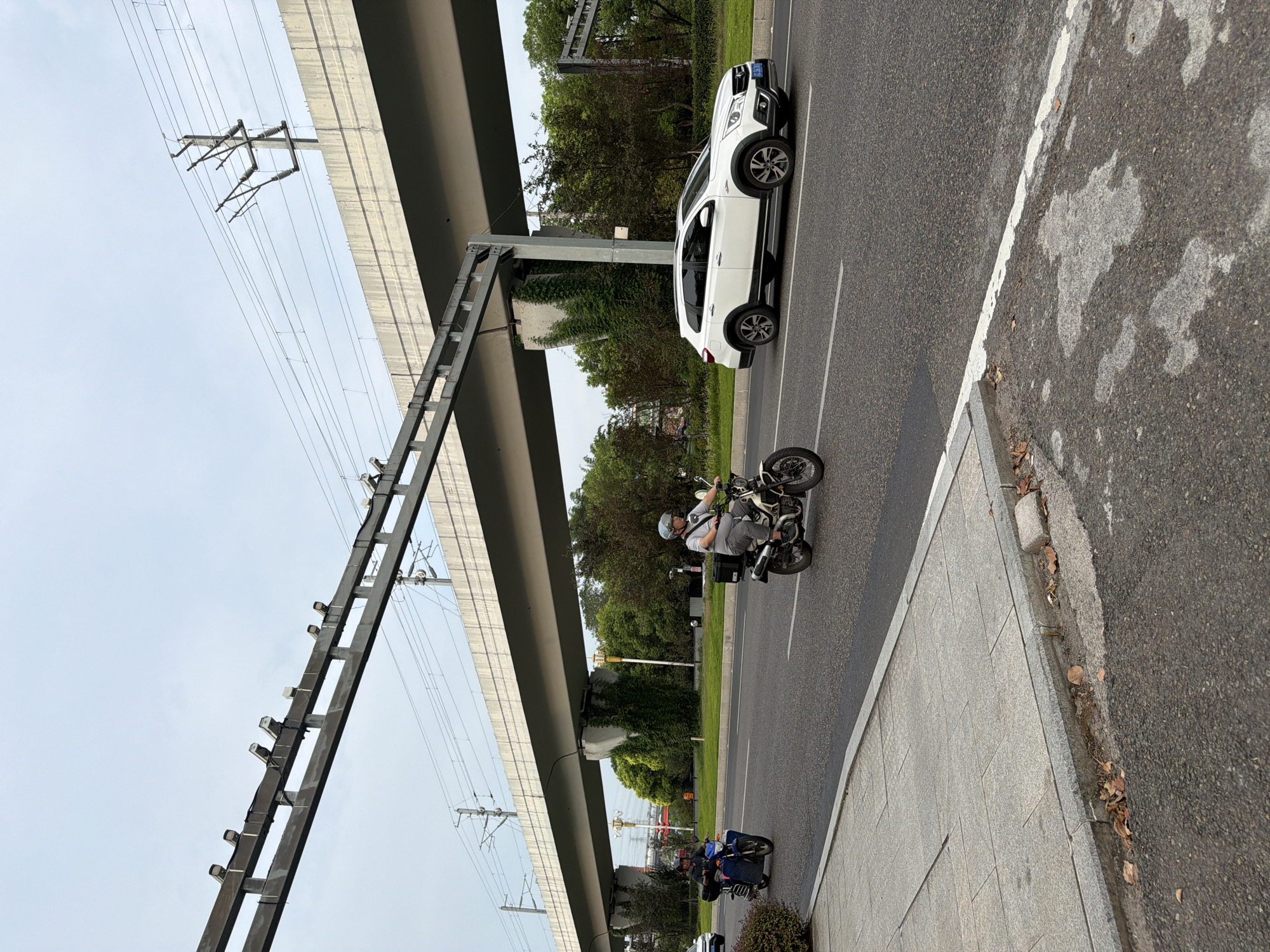 A street scene in Jiangjin, China, features an elevated overpass with overhead electrical lines and surveillance cameras. Two motorcycles and a white SUV are traveling on the asphalt road beneath the overpass. The motorcycle closest to the foreground is ridden by a person wearing a helmet and a light-colored shirt, carrying a black box on the back. A second motorcycle, ridden by a person in a helmet and dark clothing, is ahead of the first. A white SUV is in the right lane. The road has white lane markings. A concrete sidewalk with granite paving stones borders the road on the left. Trees and foliage line the sides of the road and grow on the supports of the overpass. The sky is overcast.