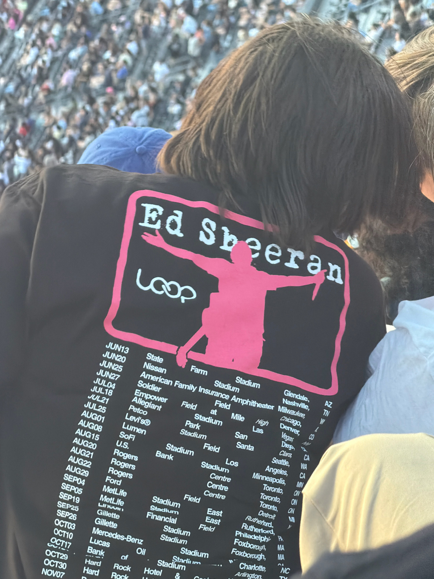 A person is seen from behind wearing a black t-shirt with a graphic of Ed Sheeran and tour dates printed in pink. The graphic displays the text "Ed Sheeran" and "Loop" with a silhouette of Ed Sheeran holding a knife. A list of tour dates and locations is printed below the graphic. The person is in a crowded outdoor venue, likely a stadium, with many other attendees visible in the background. The environment appears to be daytime, with sunlight illuminating the scene. The specific location of East Rutherford, United States of America is mentioned on the t-shirt as part of the tour itinerary.
