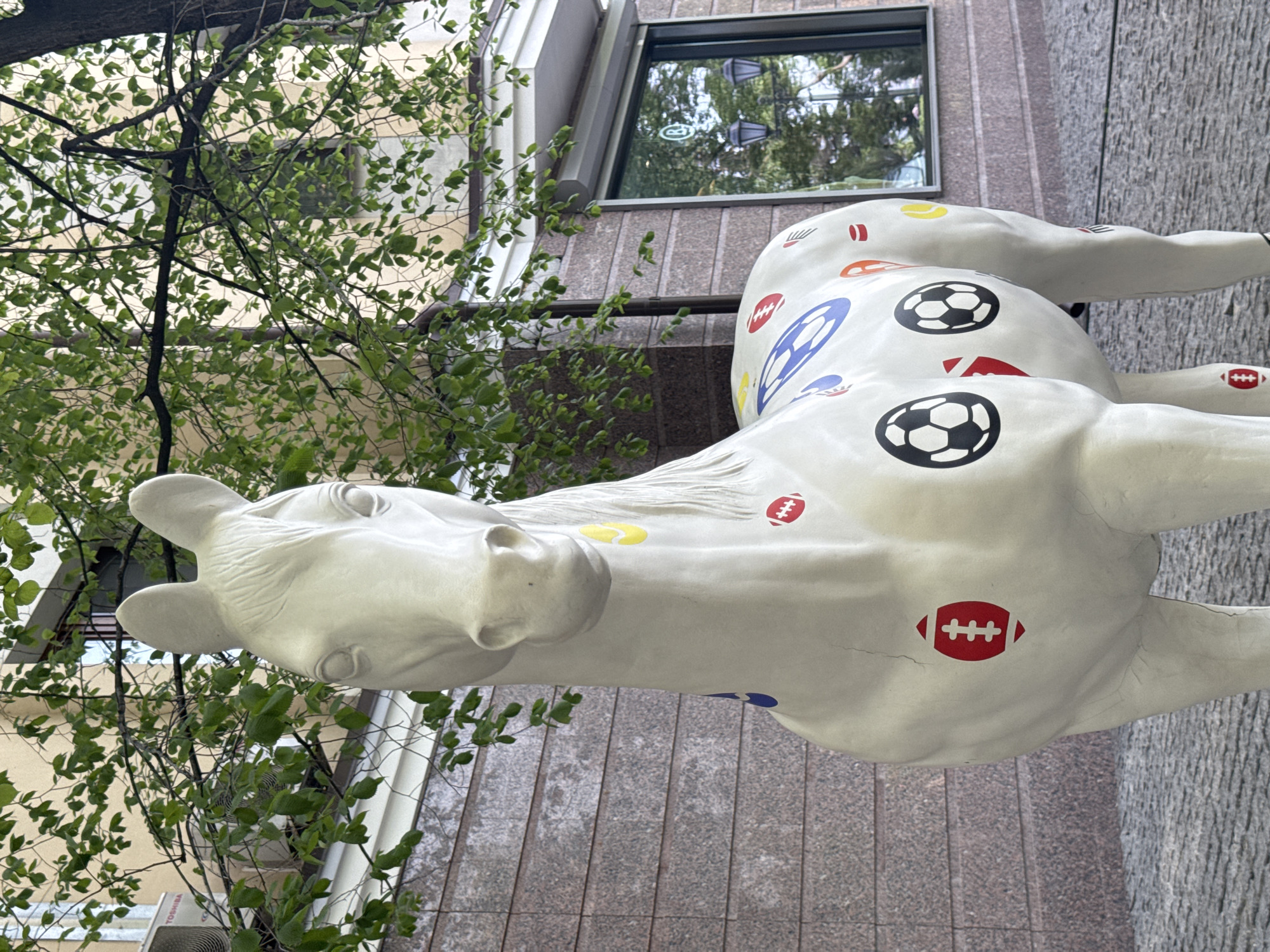 A low-angle medium shot captures a white, painted horse sculpture with sports motifs (soccer balls, footballs, etc.), standing outdoors near a building. The horse is slightly damaged. The building facade is brown, and a window is visible with reflections inside. A tree with green leaves partially obscures the left side of the image. The location is Kazan, Russia.