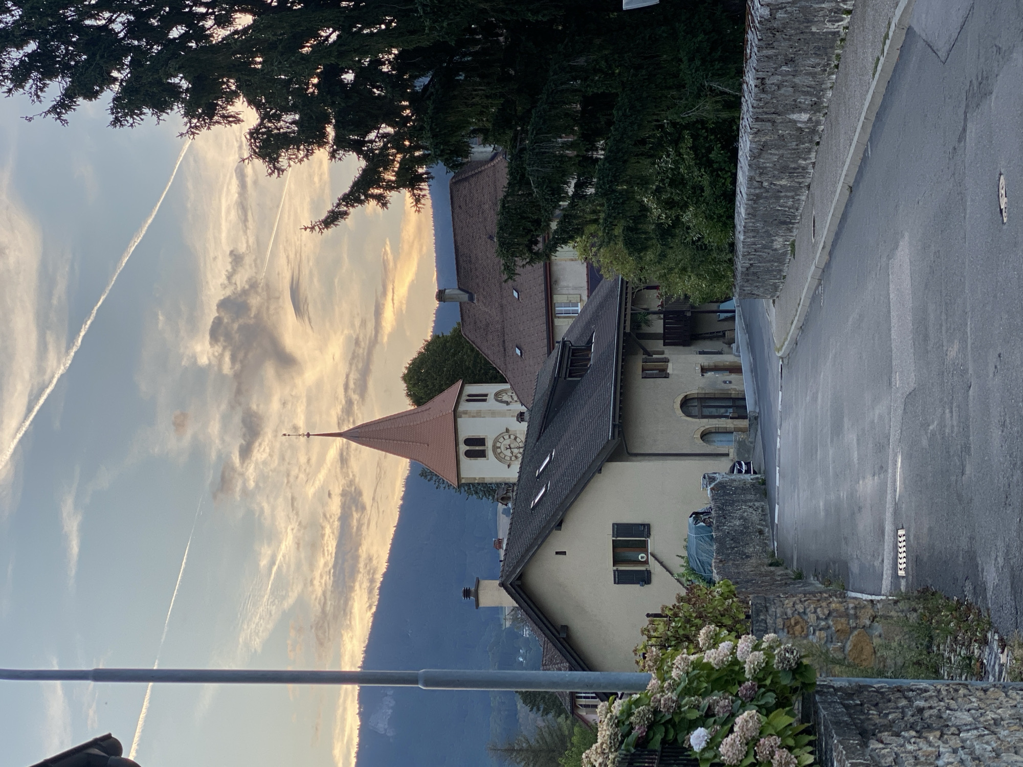 The image depicts a street scene in the city of Milvignes, Switzerland, during what appears to be late afternoon or early evening. A road curves gently upwards and to the right, bordered by stone walls. On the left, a large hydrangea bush with pink and white blooms grows beside a stone wall. Across the road, a light-colored building with a dark roof is visible, featuring shutters on a window. Further back, a church tower with a red conical roof and a clock face rises above other buildings, set against a backdrop of rolling hills and a sky with scattered clouds and contrails from aircraft. A large pine tree on the right side of the image partially obscures the view of the buildings and sky. The overall impression is of a quiet, picturesque European town.