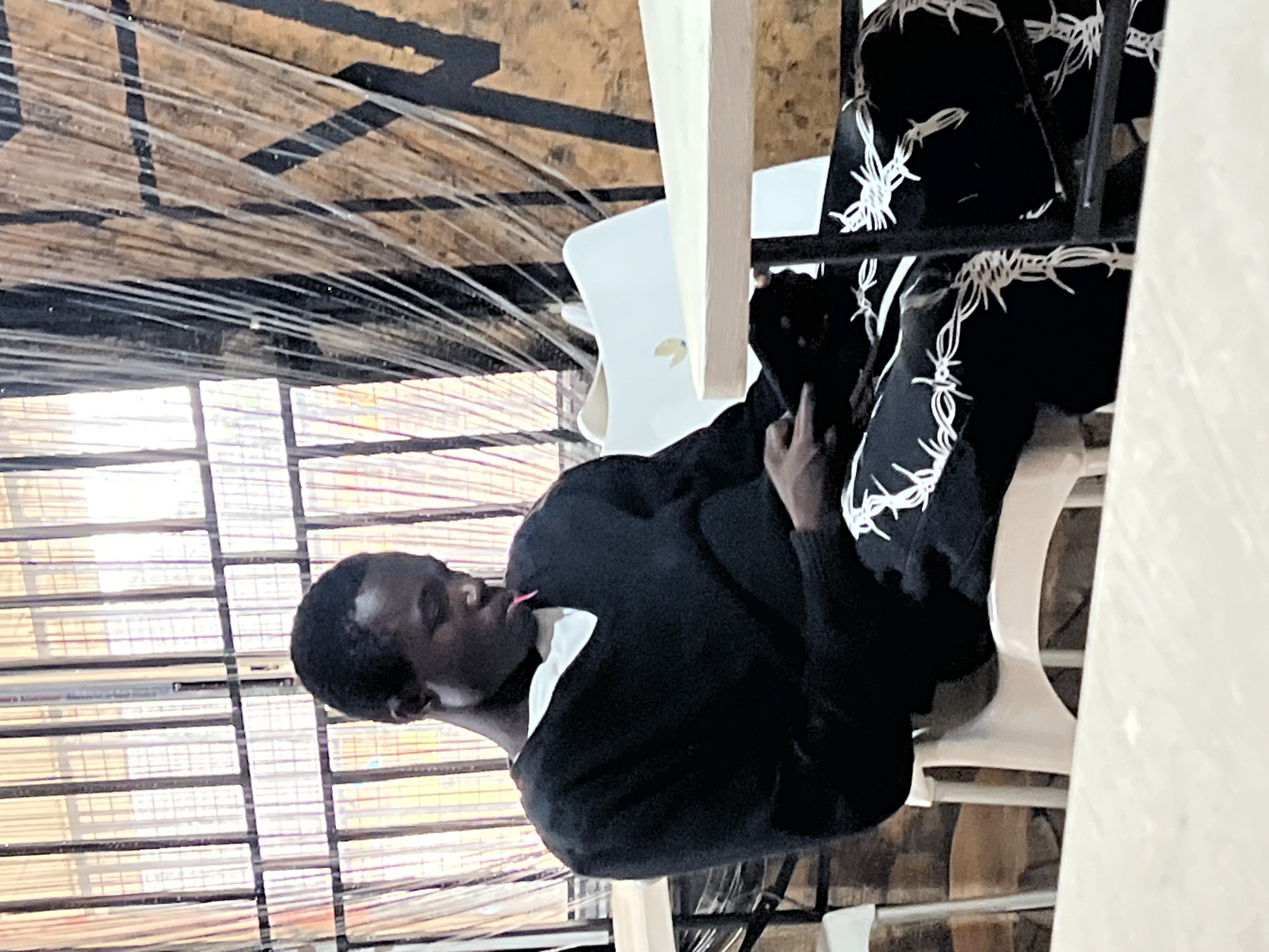 A young Black man sits at a table in what appears to be a restaurant or cafe. He is wearing a black sweater and black pants with a white barbed wire pattern. A light pink object, possibly a piece of candy, is visible between his lips. He is looking down at his hands, which are resting on his lap. The man is seated on a white plastic chair. In the background, there is a window with vertical bars, some draped fabric, and a wall with a black graphic design. Another table with a light-colored top is visible in the foreground. The scene is well-lit, suggesting it is daytime. The location is the city of Nkpor, Nigeria.