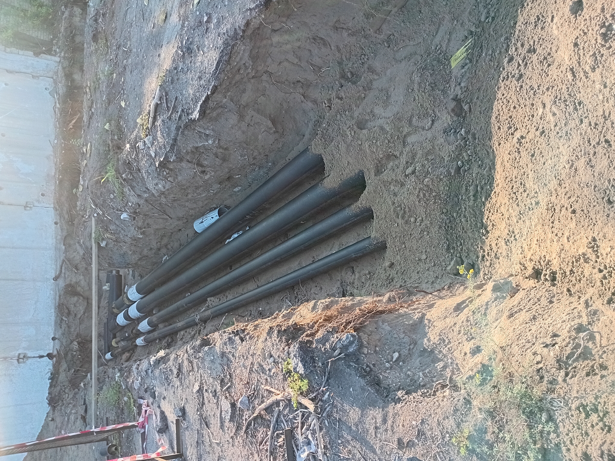 The image depicts a construction site in Semey, Kazakhstan, showing a trench dug into the earth. Several large, dark-colored pipes, likely for utilities, are laid out in the trench. The pipes are of varying diameters and appear to be made of a rigid material, possibly plastic or metal. Some of the pipes have white protective caps on their ends, and others are partially covered with soil. The trench walls are rough and uneven, revealing layers of dirt and some exposed roots. The ground surface around the trench is covered with loose soil, rocks, and debris, suggesting recent excavation work. In the background, a concrete structure or wall is visible, and there is a suggestion of greenery and dry vegetation. The lighting suggests it is daytime, possibly late afternoon, with sunlight casting shadows on the ground. There are no people visible in the image.