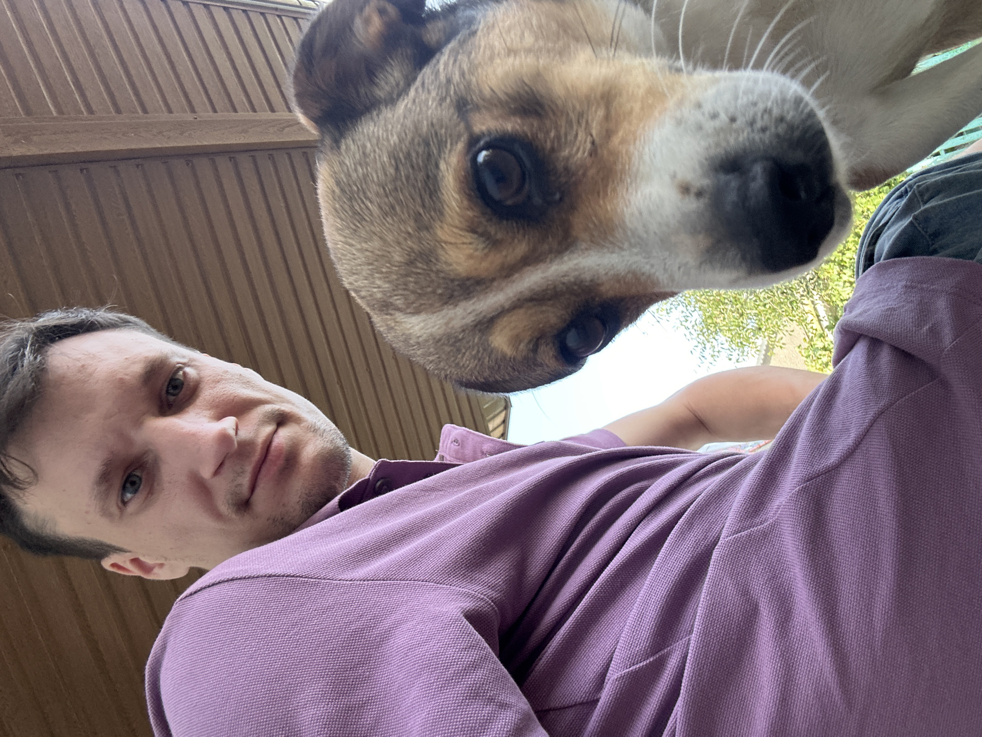 A man and a dog are shown in a close-up selfie, taken outdoors under a wooden overhang. The man, on the left, is wearing a purple polo shirt and looking towards the camera with a slight smile. His face is partially in shadow. The dog, on the right, is a medium-sized mixed breed with brown and white fur. Its face is very close to the camera, with its dark eyes and black nose prominently featured. The dog appears to be looking at the camera as well. The background shows the textured wooden ceiling of the overhang and a glimpse of green foliage and a light-colored structure, suggesting an outdoor residential area in the city of Makiv, Ukraine. The lighting indicates it is daytime.