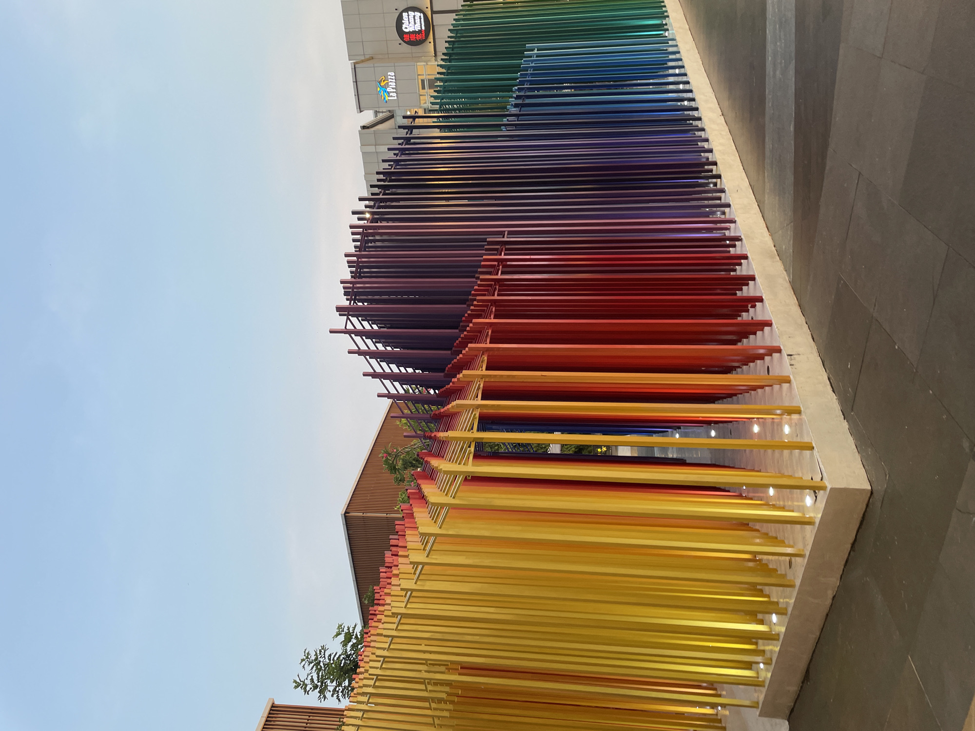 The image shows a large, colorful art installation in an outdoor setting. The installation consists of numerous parallel rods or bars, arranged in a spectrum of colors from yellow to green to blue to indigo, resembling a rainbow. The bars appear to be suspended or supported by a framework. Some lighting is integrated into the base of the structure. In the background, a building with signage including "La Pizza" and other Chinese characters can be seen. The location is speculated to be in the city of North Jakarta, Indonesia. The sky is visible in the upper portion of the image. The ground is a dark gray. There are no people visible. The perspective is from a low angle.