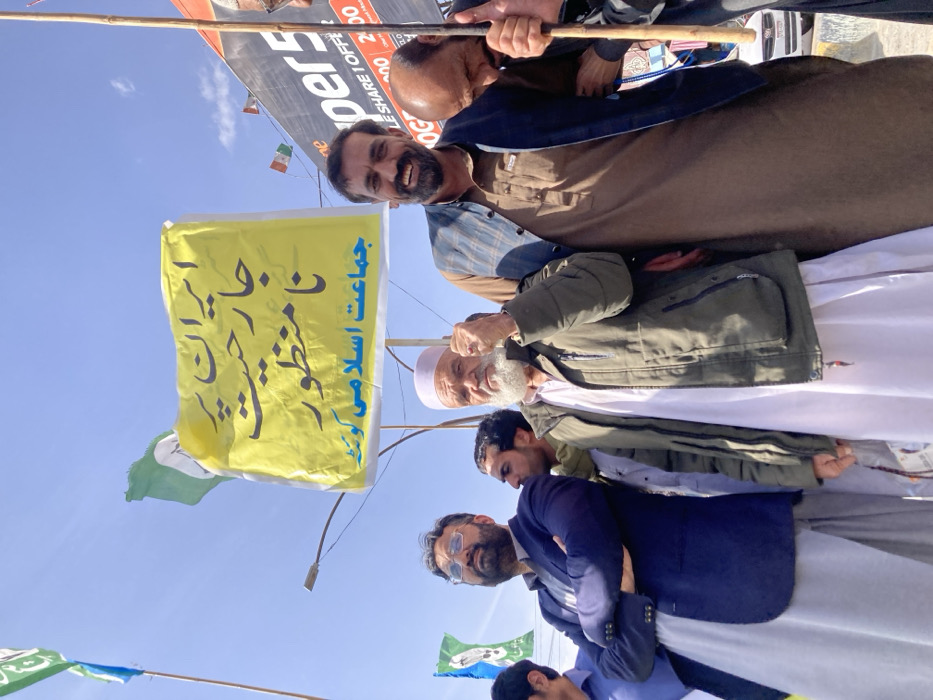 The image displays a daytime outdoor gathering of at least five men in Quetta City Tehsil, Pakistan, characteristic of a protest or political rally. In the central foreground, an older man with a prominent white beard, wearing a white shalwar kameez and an olive-green winter jacket, holds a wooden pole supporting a large yellow banner. His right hand is raised in a clenched fist. To his immediate left, a man in a dark blue blazer over a light grey shalwar kameez stands with his arms crossed, looking upwards and to the left. To the central figure's right, a man with a dark beard, dressed in a brown shalwar kameez and a dark blue sleeveless jacket, smiles directly at the camera, also appearing to hold a banner pole.
The prominent yellow banner displays black Urdu script. The top line translates to "Aggression against Iran is unacceptable," and the bottom line identifies the organizing body as "Jamaat-e-Islami Quetta."
In the background, multiple green and white flags featuring a weighing scale symbol, indicative of Jamaat-e-Islami, are visible on poles, predominantly on the left side of the frame. Further back on the right, small flags with green, white, and orange horizontal stripes are present. A large commercial billboard displaying "super5" and partial pricing information is visible on the far right. The sky overhead is clear and blue, with minimal cloud cover. A street light pole is also visible behind the banner. A white vehicle is partially discernible in the far background on the right.