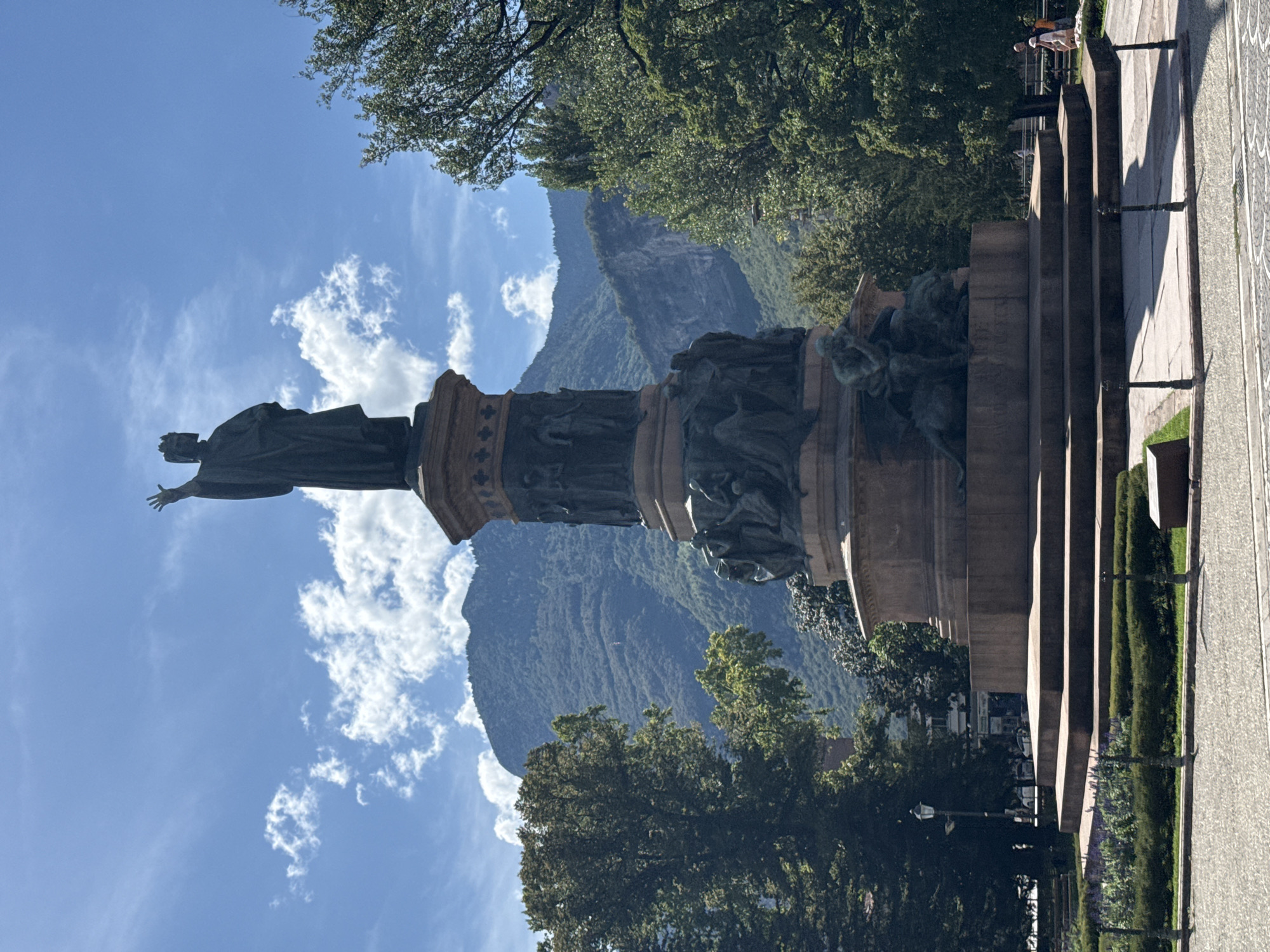 The image shows a statue of a crowned figure with an outstretched arm, standing on a tall, ornate pedestal. The statue is located in a park-like setting with trees and manicured hedges. In the background, a large, forested mountain range is visible under a blue sky with scattered clouds. The base of the statue features several bronze figures, including one that appears to be a winged creature. In the lower right corner, two people are walking on a paved path. A lamppost is visible to the left of the statue, and buildings can be seen behind the trees. The foreground consists of a paved area and a low fence. The statue is likely the Monumento a Dante in Trento, Italy, given its appearance and the mountainous backdrop characteristic of the region.