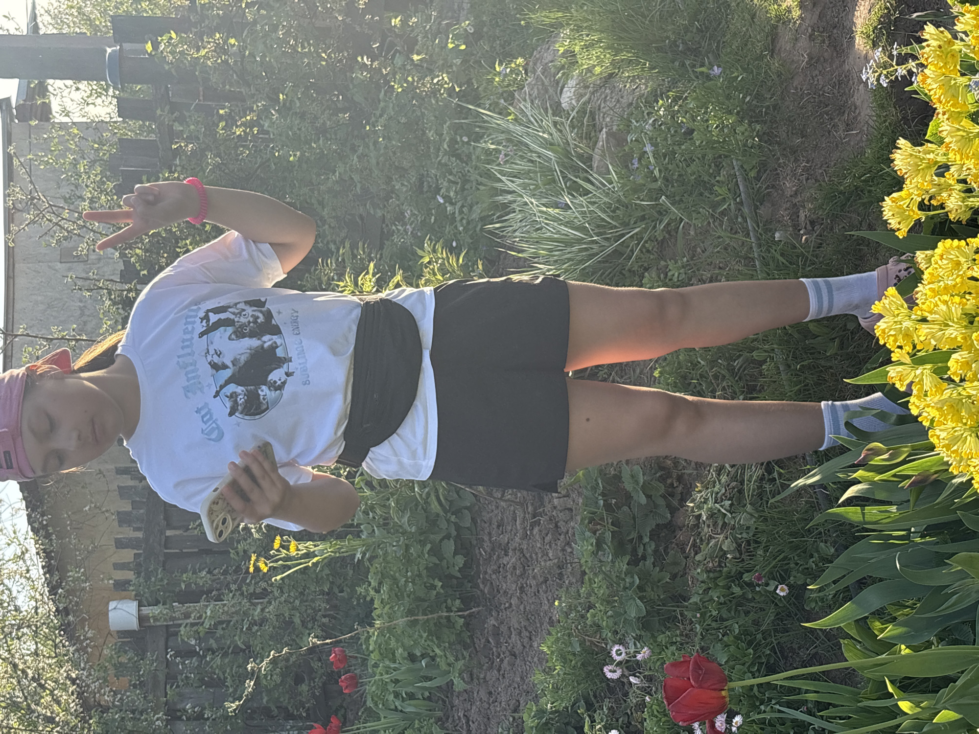 A girl wearing a pink headband, a white t-shirt, and black shorts stands in a garden. She is holding a phone in her right hand and making a peace sign with her left hand. She is wearing white socks with blue stripes. The garden contains red tulips, yellow primroses, daisies, and various green plants. A wooden fence is visible in the background. This photograph likely took place in Chaikovsky, Russia.