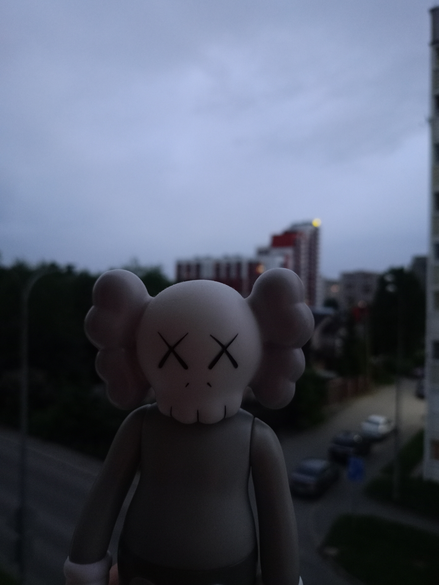 The image shows a light gray figurine with cartoonish "X" eyes, reminiscent of KAWS art, positioned in the foreground. The figurine is facing forward, and its body is mostly visible. In the background, a city street with parked cars is visible, indicating an urban setting. Several multi-story buildings are also present, some with red accents. The sky is overcast and appears to be either early morning or late evening. The scene is likely Petrozavodsk, Russia, given the context provided.