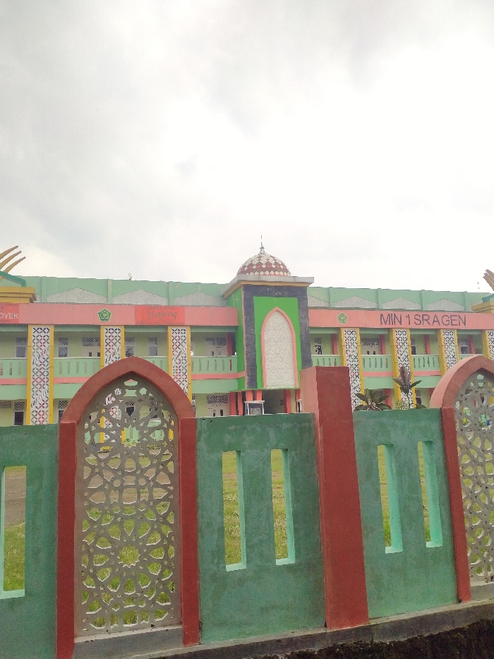 An exterior view captures a colorful building, likely an educational institution, seen from a slightly low angle through a decorative fence. The building features a multi-story structure painted predominantly in light green and pink, with accents of orange, yellow, and purple. Its architecture incorporates Islamic design elements, including a central facade with a prominent arched recess and a small, patterned dome on the roofline. Geometric patterns adorn the building's walls and arches, particularly on the second floor and around windows.

In the foreground, a green and red fence runs across the bottom of the frame. This fence has several sections: a decorative arched opening with an intricate white geometric pattern on the left, and solid green panels with rectangular cut-outs on the right, separated by red pillars. Through the fence, a patch of green grass is visible.

The sky above is bright and overcast, suggesting daytime with cloudy weather.

Visible text on the building includes "MIN 1 SRAGEN" clearly displayed on the right side of the facade, indicating it is Madrasah Ibtidaiyah Negeri 1 Sragen, an Islamic elementary school. Other partial text elements, "Malang" and "OYEK," are also visible on the left side of the building. The vibrant colors and distinct architectural style contribute to a cheerful and inviting appearance of the school, located in Tanon, Indonesia. No people or activities are visible in the image.