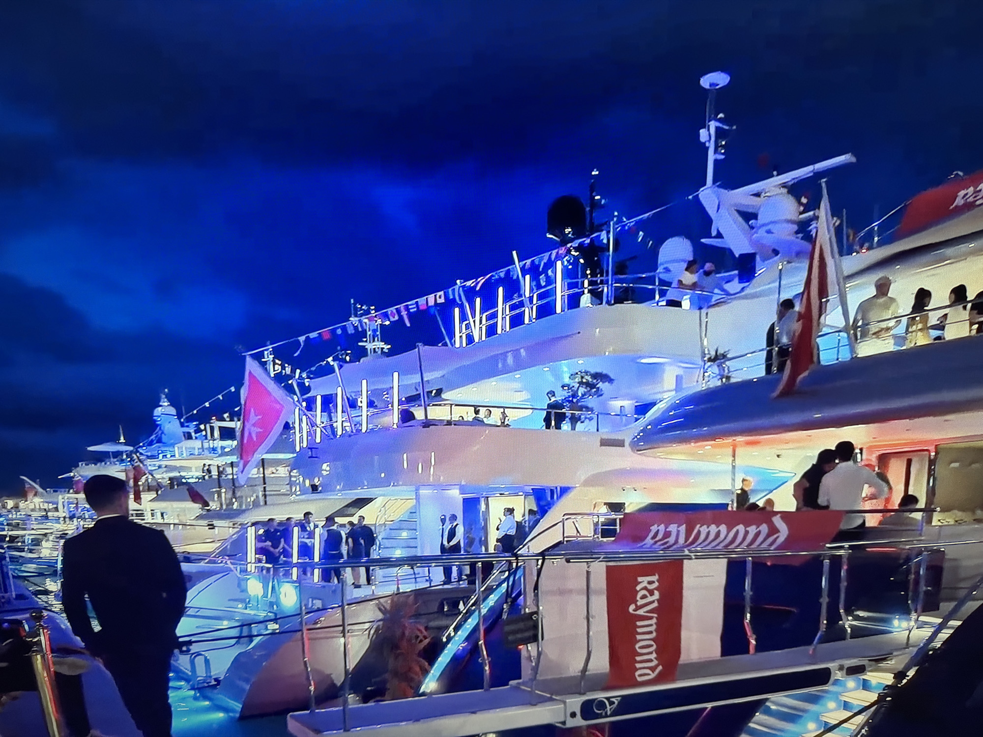 A nighttime aerial shot captures multiple luxury yachts docked in the city of Okha, Russia. The yachts are illuminated with vibrant blue and white lights, casting a colorful glow on the dark water. Many of the yachts are adorned with decorative flags and bunting, adding to the festive atmosphere. Several individuals are visible on the decks of the yachts, some appearing to be conversing while others stand or move about. One prominent yacht features a large red banner with white lettering that spells out "Raymond." The overall scene suggests a gathering or event taking place on the water, possibly a celebration or exhibition of the yachts.