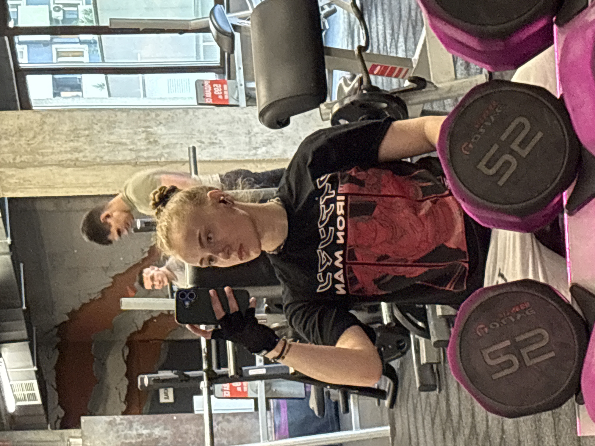 A woman is taking a selfie in a gym. She is wearing a black Iron Maiden t-shirt and fingerless gloves. She is holding a black smartphone. A man is visible in the background. Weights are in the foreground, specifically 52lb dumbbells. The gym has a window and what appears to be a concrete or tiled wall. The exact location in Sofia, Bulgaria is not discernible from the image.