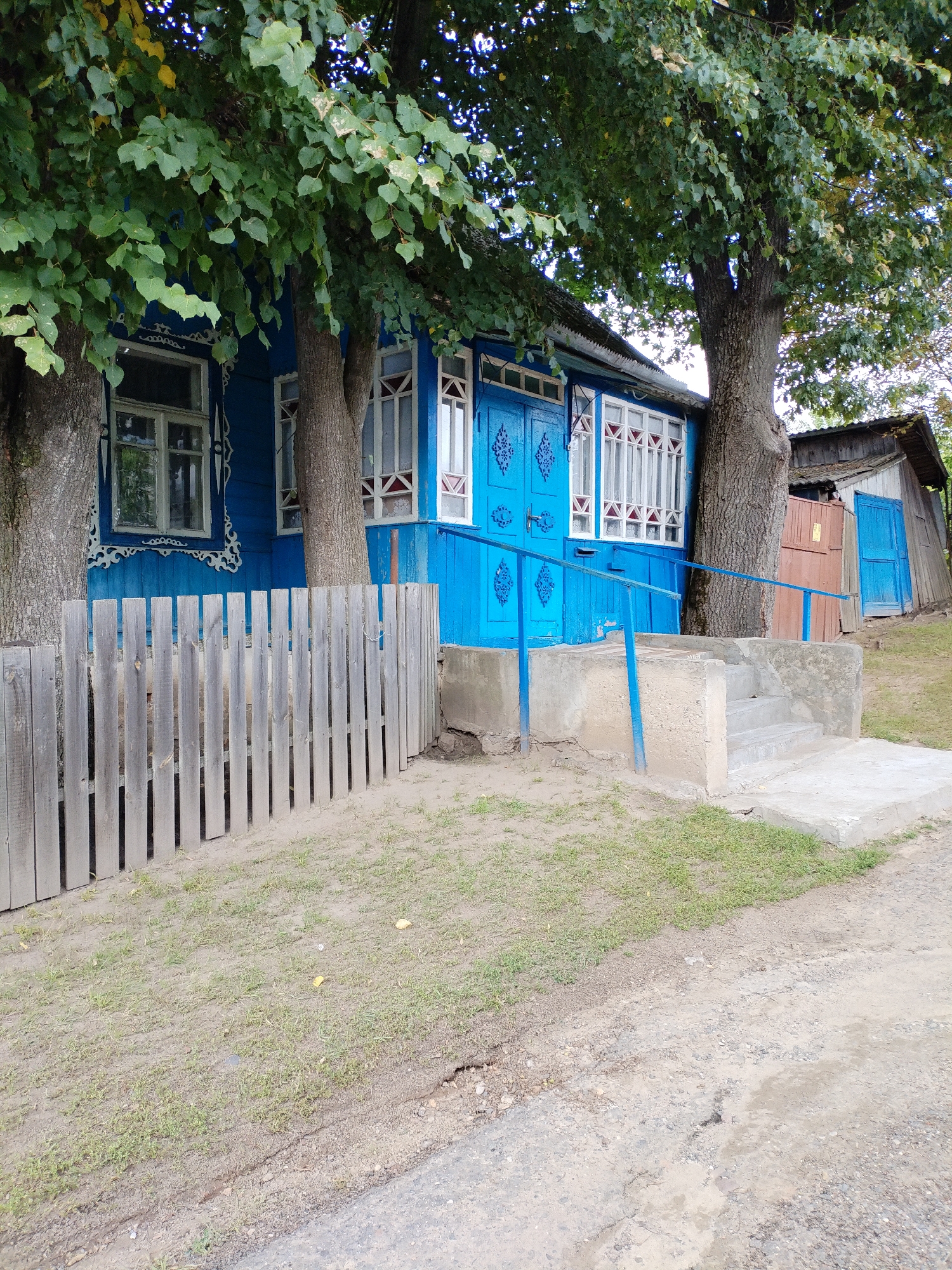The image depicts a blue wooden house with white-trimmed windows and ornate carvings around them. Two large trees with green foliage partially obscure the house. A wooden fence runs along the left side of the frame, and a concrete staircase leads to the house's entrance, which features blue double doors. To the right of the house, a smaller wooden shed with a blue door is visible. The ground in front of the house is a mix of dirt and sparse grass, with a dirt road in the foreground. The scene appears to be in a rural or suburban setting in Bialynichy, Belarus, given the architectural style and the presence of trees and a modest dwelling. There are no people or animals present in the image, and no specific actions are depicted. The overall impression is one of a simple, lived-in dwelling in a natural environment.