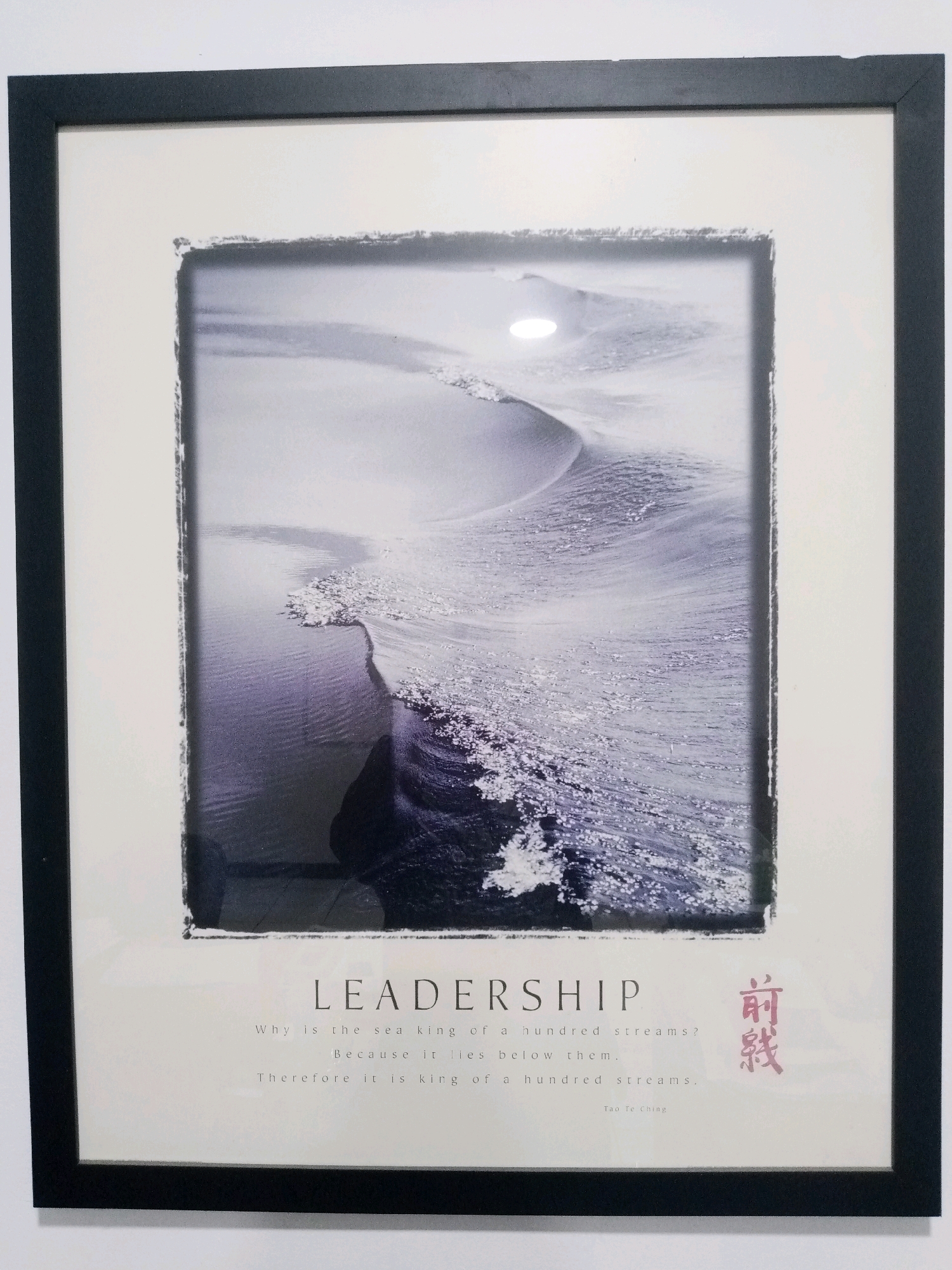 The image displays a framed motivational print hanging on a white wall. The print features a black and white photograph of a wave breaking on a shore, with a spotlight reflecting on the water. Below the photograph, the word "LEADERSHIP" is printed in large, bold letters. Beneath this, a quote is presented in smaller text: "Why is the sea king of a hundred streams? Because it lies below them. Therefore it is king of a hundred streams." The quote is attributed to "Tao Te Ching." To the right of the quote, there is a red-ink stamp with Chinese characters. The frame is black and has a matte white border around the print. There is no indication of the location within the city of Balaraja, Indonesia.