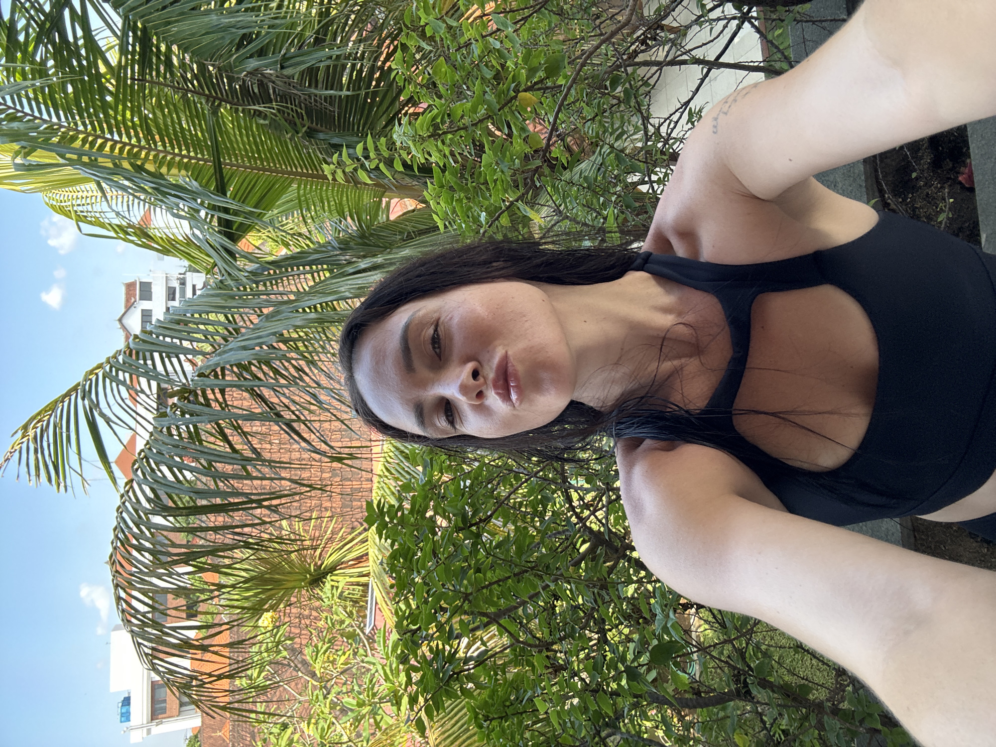 A woman is taking a selfie outdoors in North Kuta, Indonesia, with lush green foliage and palm trees in the background, along with buildings. The woman has dark hair, is wearing a black athletic top, and is puckering her lips. The lighting suggests it is daytime.