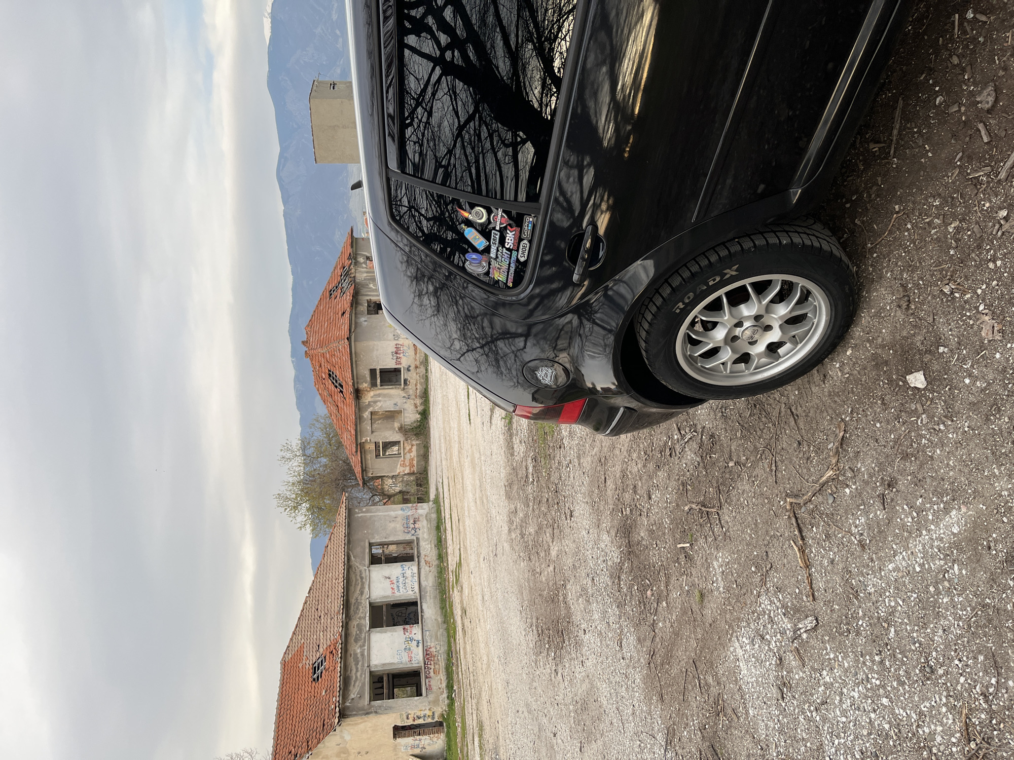 The image shows a black car parked on a gravel surface in front of dilapidated buildings with damaged roofs. Graffiti is visible on the buildings. The car is oriented toward the right of the frame. The rear left tire of the car is prominently displayed. Mountains are visible in the background, obscured partially by clouds. Based on the buildings and the location, it is plausible that the scene is near the abandoned buildings in Karavelovo, Bulgaria. There are no people in the frame.