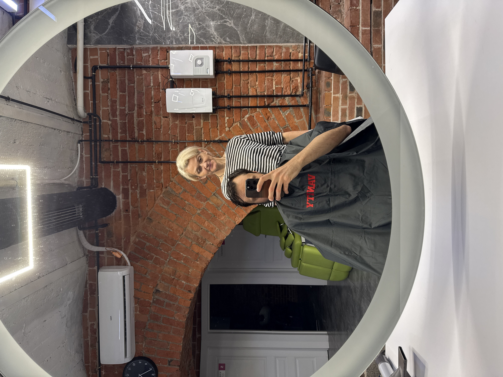 This image captures a mirror selfie taken inside what appears to be a modern hair salon or barbershop in Moscow, Russia. The shot is a reflection in a large, circular mirror with an illuminated edge, positioned above a white counter.
In the reflection, a blonde woman, likely a stylist, stands to the right of a seated male client. She is smiling warmly at the camera (the client's smartphone), and wears a black and white striped top. The client, with dark hair, is mostly obscured by his phone, which he holds up to take the picture, effectively capturing this mirror shot. He is covered by a dark gray protective cape with red text that reads "YAMAY" (appearing reversed as "YT MAY" in the reflection). A bright green upholstered chair is visible behind him in the background.
The interior design features a striking industrial aesthetic with exposed red brick walls and visible black pipes, particularly prominent in the upper background of the reflected image. An arched brick opening is visible towards the left side of the reflection. On the left wall, outside the mirror's frame, a white Haier air conditioner unit is mounted, with a round black clock visible below it. Further to the right, on another brick wall, two white rectangular units, possibly water heaters or control panels, are mounted, connected by black piping. Some grey textured wall elements and white concrete sections also contribute to the background.
The atmosphere appears friendly and relaxed, indicated by the stylist's smile. The time of day is not evident, as the scene is brightly lit by interior lighting, including the mirror's illumination.
Visible text includes "YAMAY" on the cape, "Haier" on the air conditioner, and numbers on the clock (11, 12, 1, 2, 3). Some faint Russian text "2 KVARTIR" is partially visible in the reflection near the bottom right, possibly from a door sign.