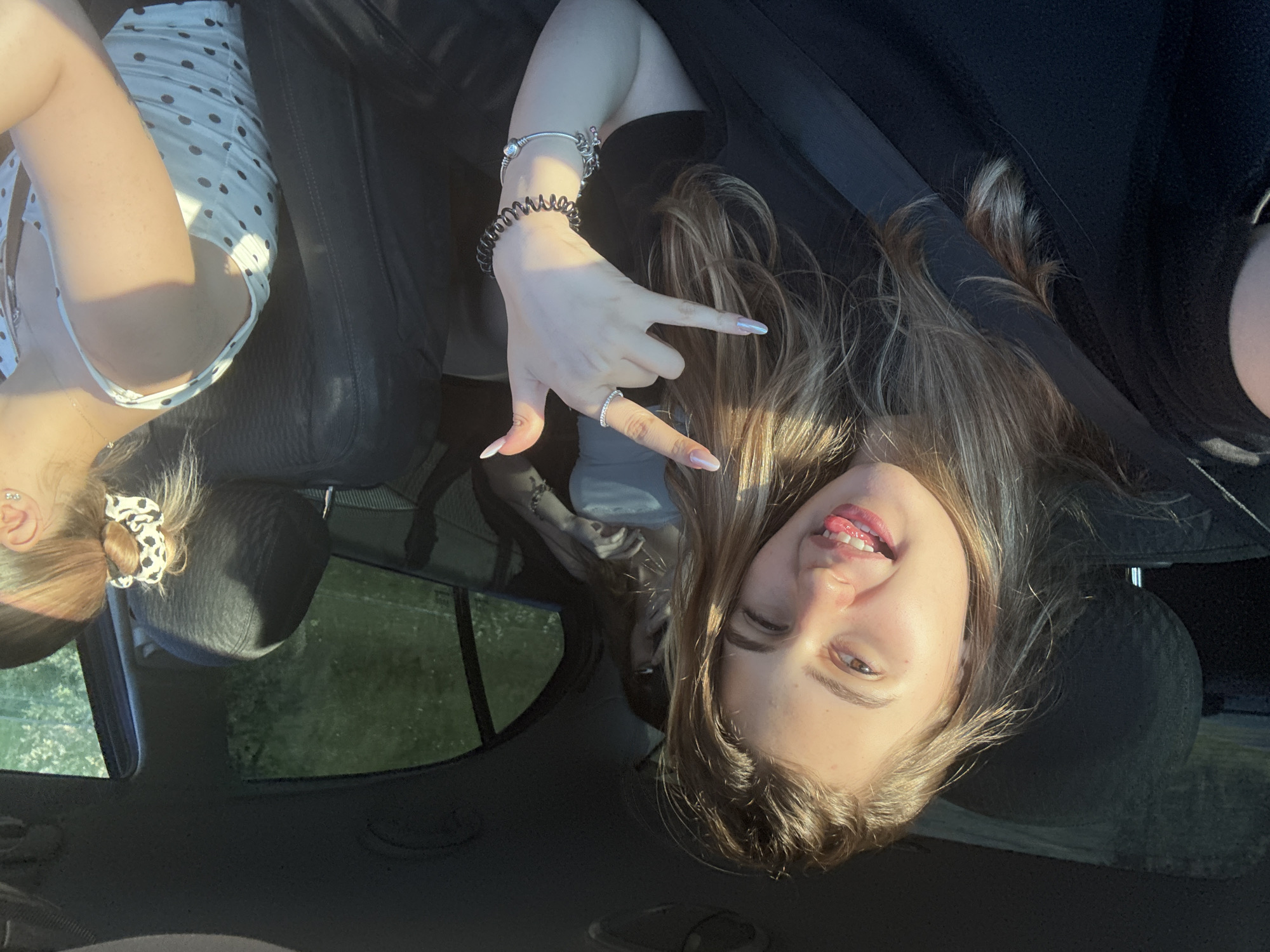 This is a selfie taken by a young woman inside a car. She is in the passenger seat, and her head is tilted back. She has long, blonde hair and is wearing a black t-shirt. Her left hand is raised, and she is making a "rock on" gesture with two fingers. She is also wearing a ring on her left ring finger and several bracelets on her left wrist. Her tongue is sticking out of her mouth, and she appears to be smiling.
Another person is visible in the background, in the driver's seat. This person is wearing a white dress with black polka dots. They are also looking towards the camera.
The scene appears to be outdoors, with greenery visible through the car windows. The lighting suggests it is daytime, possibly late afternoon, given the warm glow. The interior of the car is dark, with black leather seats. The overall mood of the image is playful and fun.
There is no visible text in the image. The location context is Koprivshtitsa, Bulgaria, which is a historic town known for its well-preserved Bulgarian Revival architecture and its role in the Bulgarian National Revival.
