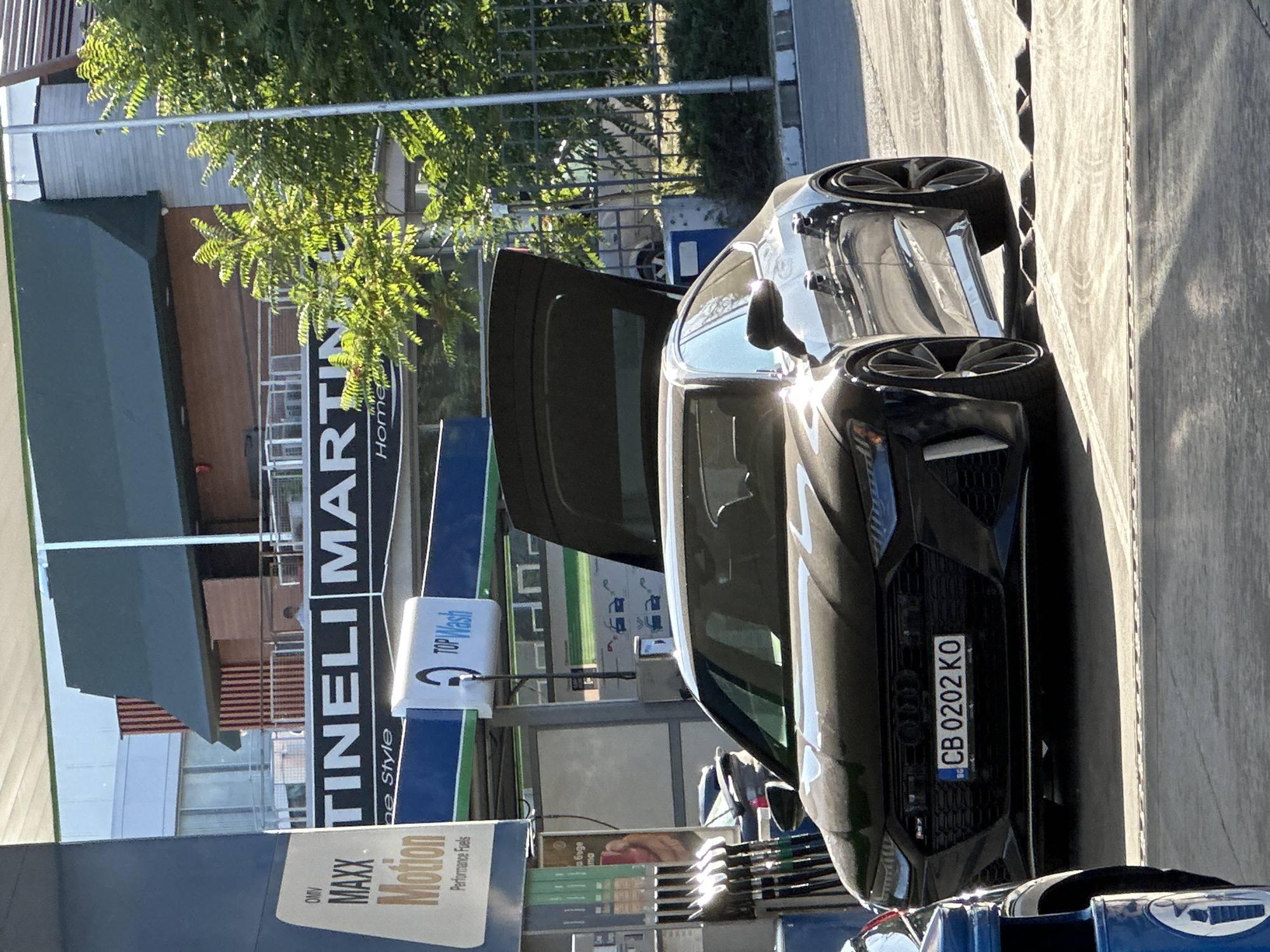 A black Audi RS7 with an open trunk is parked at a gas station. The license plate reads "CB 0202 KO". The car is positioned in front of a gas pump. In the background, there is a building with signage that includes "MARTINI". The scene appears to be a sunny day. This gas station is located in the city of Veliko Tarnovo, Bulgaria.