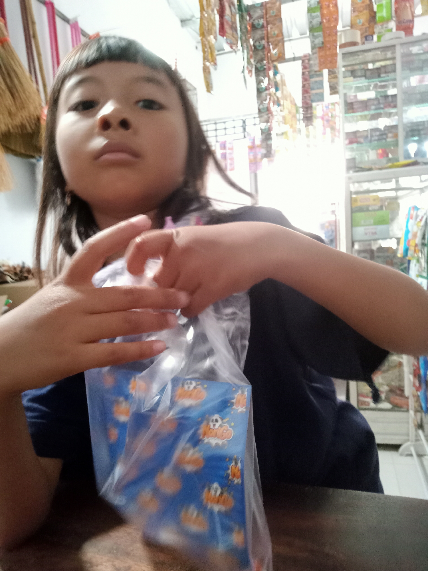 The image shows a young child, likely of Asian ethnicity, in the foreground, facing the camera. The child has dark hair styled with bangs and is wearing a dark blue shirt. They are holding a clear plastic bag containing what appears to be candy or snacks with a blue wrapper featuring orange pumpkin-like shapes and cartoonish figures. The child's hands are engaged in opening or manipulating the bag.
The background suggests an indoor environment, possibly a small shop or convenience store. Shelves filled with packaged goods, snacks, and various items are visible, with some items hanging from the ceiling. The lighting is bright, with some areas appearing overexposed. The overall setting is consistent with a retail space in Simo, Indonesia. A dark wooden surface is in the immediate foreground, indicating the child is sitting at a table or counter.