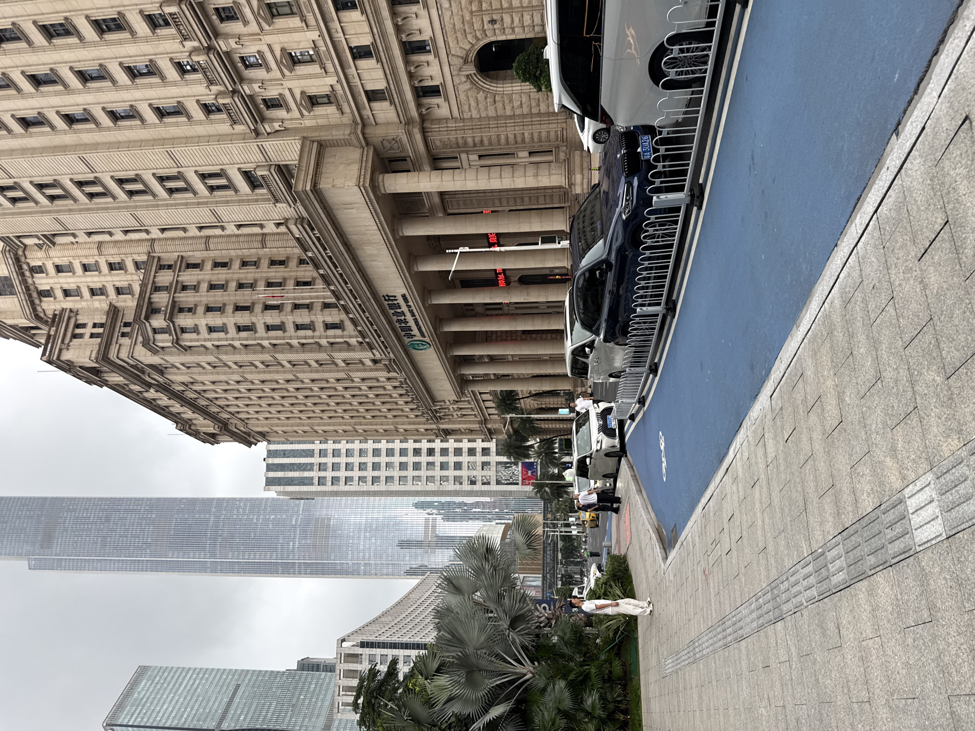 This image depicts an urban scene in Tianhe District, China. The setting is a street with a large, ornate building on the left, characterized by its stone facade, numerous windows, and classical columns. In front of this building, several cars are parked along a blue-colored pathway. The pathway is bordered by a metal fence. Further down the street, modern skyscrapers rise into a cloudy sky.
In the foreground, a gray tiled sidewalk is visible, with a tactile paving strip for visually impaired pedestrians running along its edge. A person dressed in white is standing on the sidewalk, looking towards the buildings. To the right of the sidewalk, there are lush green plants and palm trees.
The image appears to have been taken during the daytime, with overcast weather conditions indicated by the diffused light and the gray sky. The overall atmosphere is that of a bustling urban environment with a mix of historical and modern architecture.
On the ornate building, some Chinese characters are visible, which appear to be part of a sign for a bank. The license plate of a parked car shows "31126". There is also a red sign with some text, but it is not clearly legible.