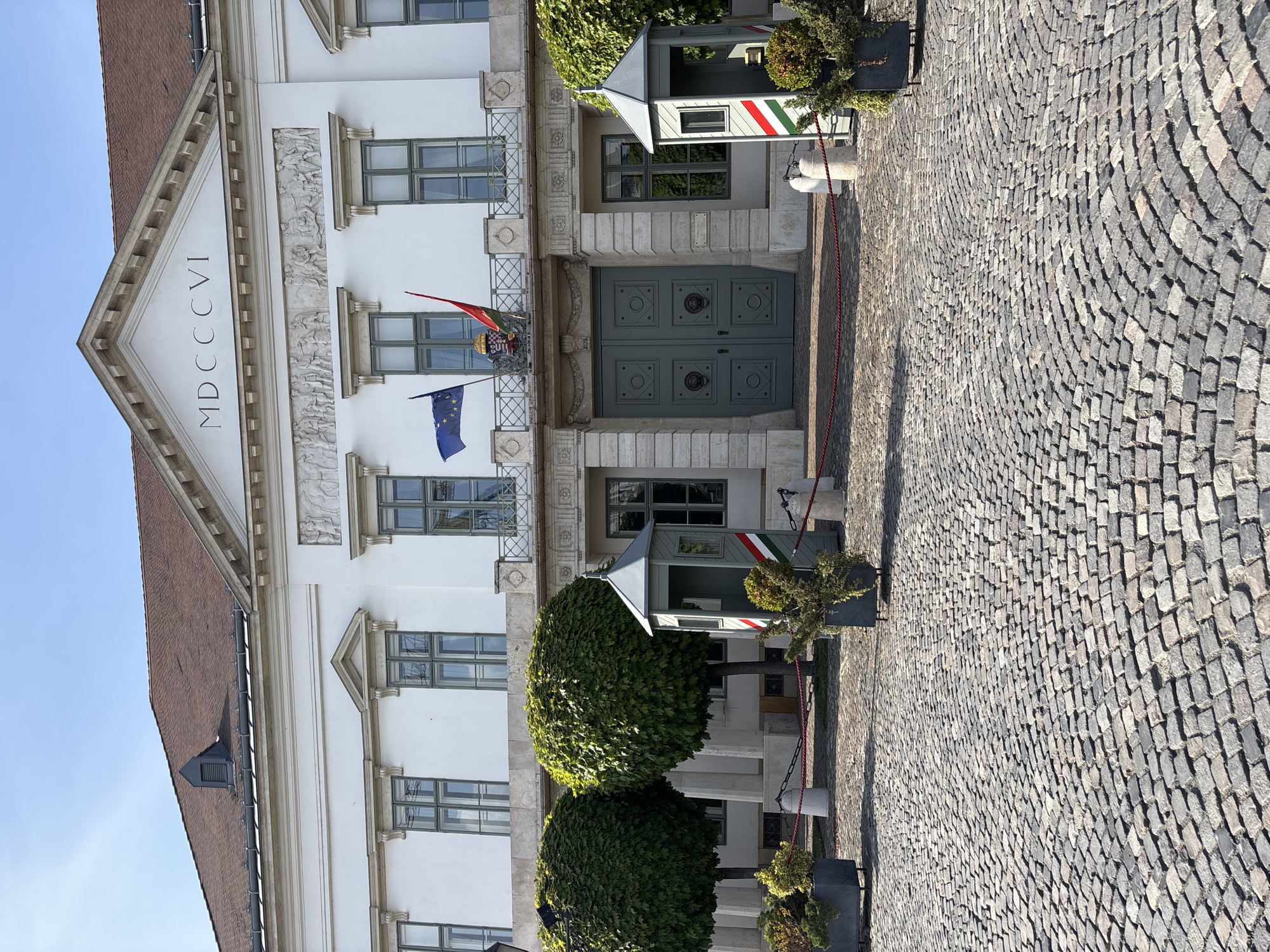 A well-maintained white building with classical architectural elements stands under a clear blue sky on a bright, sunny day. The building features several windows with green frames and a prominent central entrance with a large, ornate green double door. Above the entrance, a European Union flag and a Hungarian flag, complete with a crest, are displayed. The facade is adorned with decorative bas-reliefs and pilasters. On the triangular pediment at the top of the building, the Roman numerals "MDCCCVI" are inscribed.
On either side of the entrance area, two guard booths (sentry boxes) are visible, painted with the distinctive red, white, and green stripes of the Hungarian flag. Red rope barriers stretch between stone posts, delineating a path or secure area. The ground leading up to the building is covered in an intricate pattern of gray cobblestones. Lush, spherical green bushes and smaller potted plants are strategically placed, adding natural elements to the formal setting. No people are visible, and no specific event is taking place beyond the general view of a stately, possibly official, building. The presence of the Hungarian flag colors on the guard booths strongly suggests a location in Hungary.