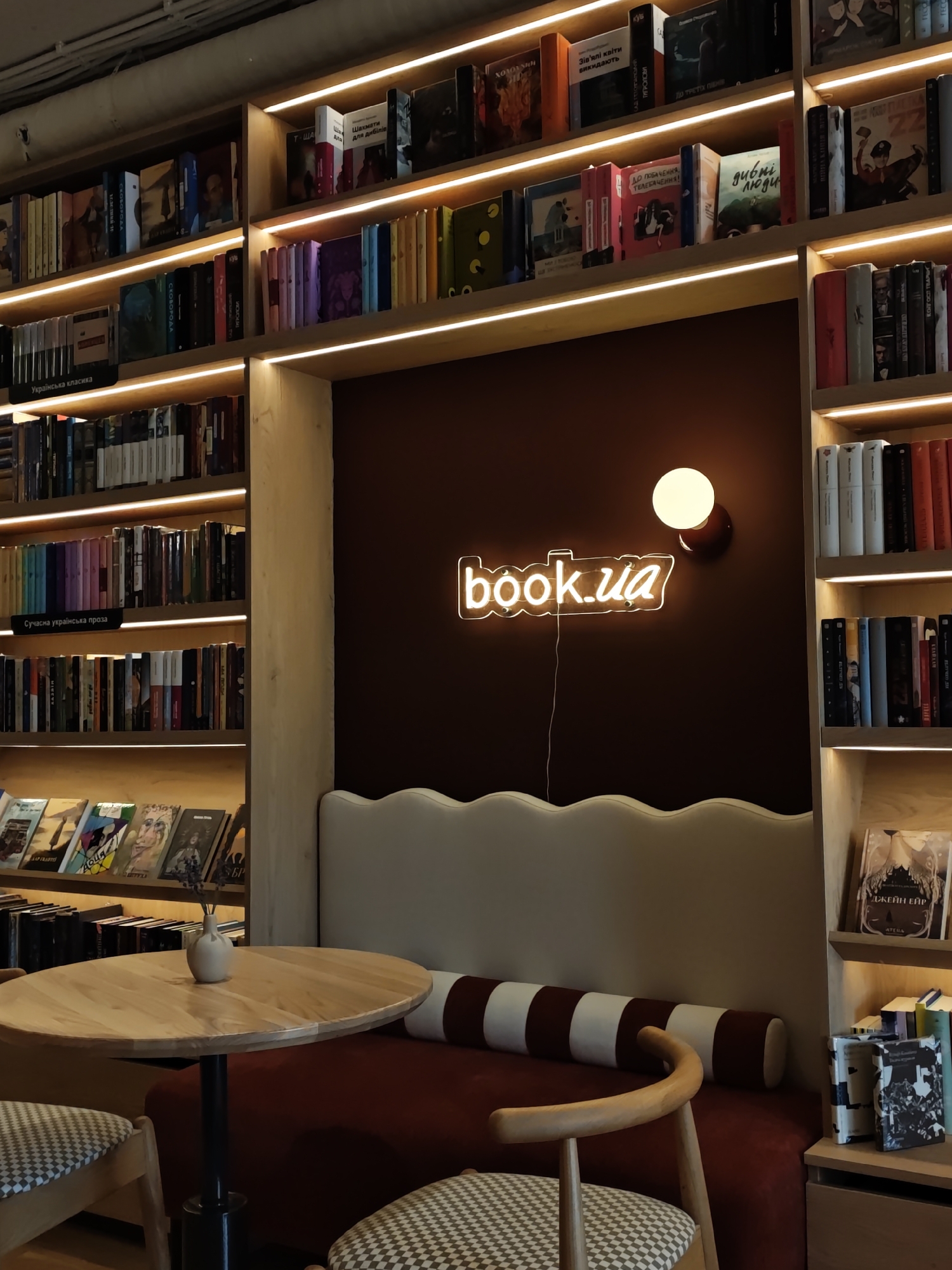 The image depicts the interior of a bookstore, likely in Kyiv, Ukraine, given the "book.ua" neon sign, which is a popular Ukrainian online bookstore. The scene features floor-to-ceiling bookshelves filled with books, illuminated by warm LED strip lighting along each shelf. In the foreground, a round wooden table with a small vase of dried flowers sits between two chairs with patterned upholstery. Behind the table and chairs is a dark red, cushioned bench with a wavy backrest, also illuminated by soft lighting. A prominent neon sign on the dark brown wall reads "book.ua" in bright white letters. The overall ambiance suggests a cozy and inviting reading or browsing space within a bookstore.