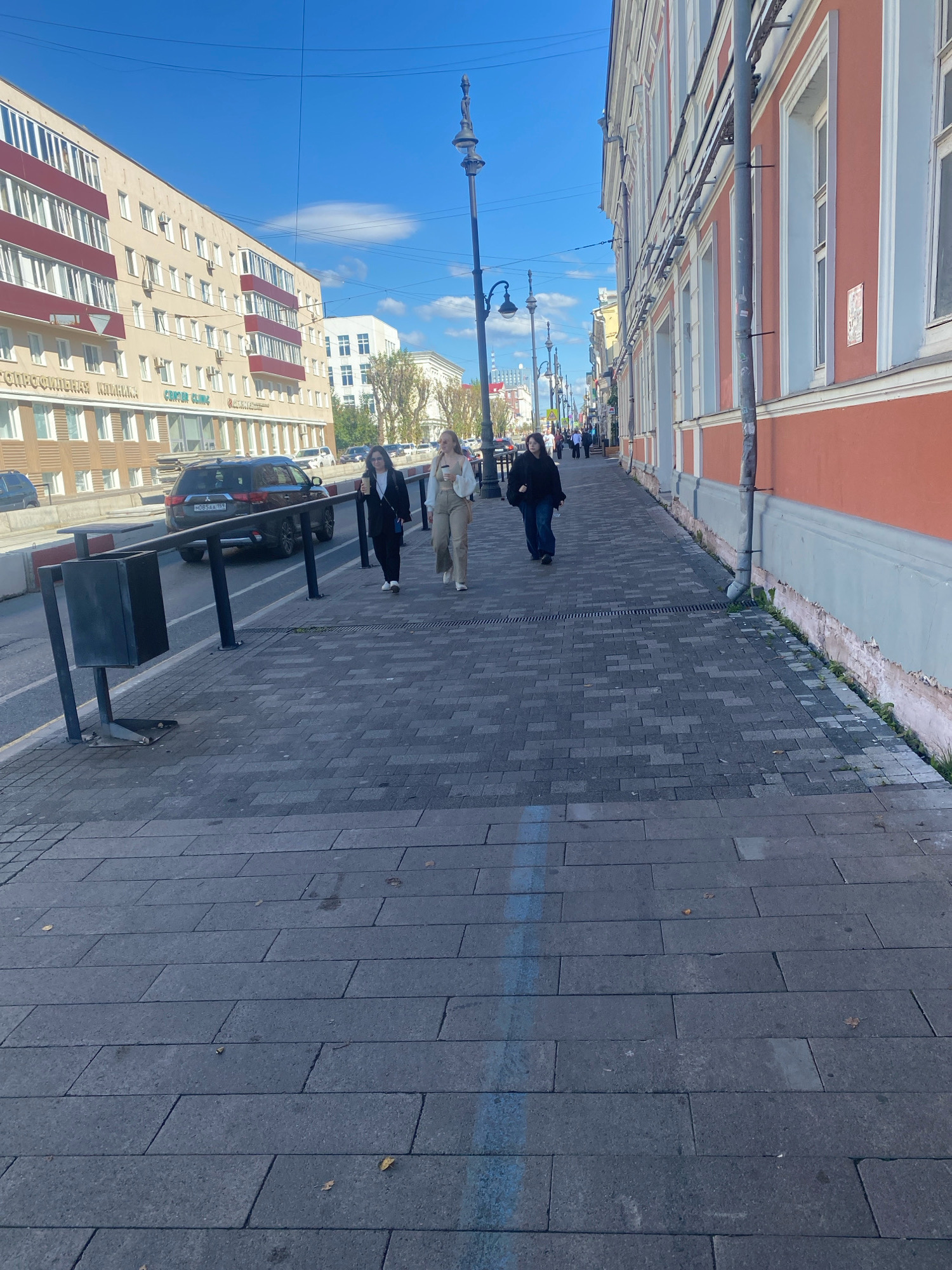 A street scene in Perm, Russia, features a wide pedestrian sidewalk paved with dark grey rectangular stones, laid in a staggered pattern. The sidewalk runs alongside a street with a dark-colored SUV driving on it.
On the sidewalk, three individuals are walking away from the camera. The person in the foreground is wearing a dark suit jacket and pants, with white sneakers, and carrying a light-colored disposable cup. To their right and slightly ahead, another person is wearing a beige outfit, consisting of trousers and a light-colored jacket, also carrying a disposable cup. Further ahead and to the left, a person in a black jacket and jeans walks with their head down.
To the left of the sidewalk is the street, with a dark SUV bearing the license plate "M085AA 159 RUS". Beside the street, there is a dark metal trash receptacle on a black metal stand. In the background, to the left, a multi-story apartment building with beige facades and maroon balconies stands under a clear blue sky with scattered white clouds. To the right of the street, a building with an orange facade and white trim lines the sidewalk. The base of this building shows signs of wear and peeling paint, with small plants growing along the foundation. Streetlights with traditional designs are visible along the sidewalk. The overall environment suggests a moderately urban setting on a sunny day.