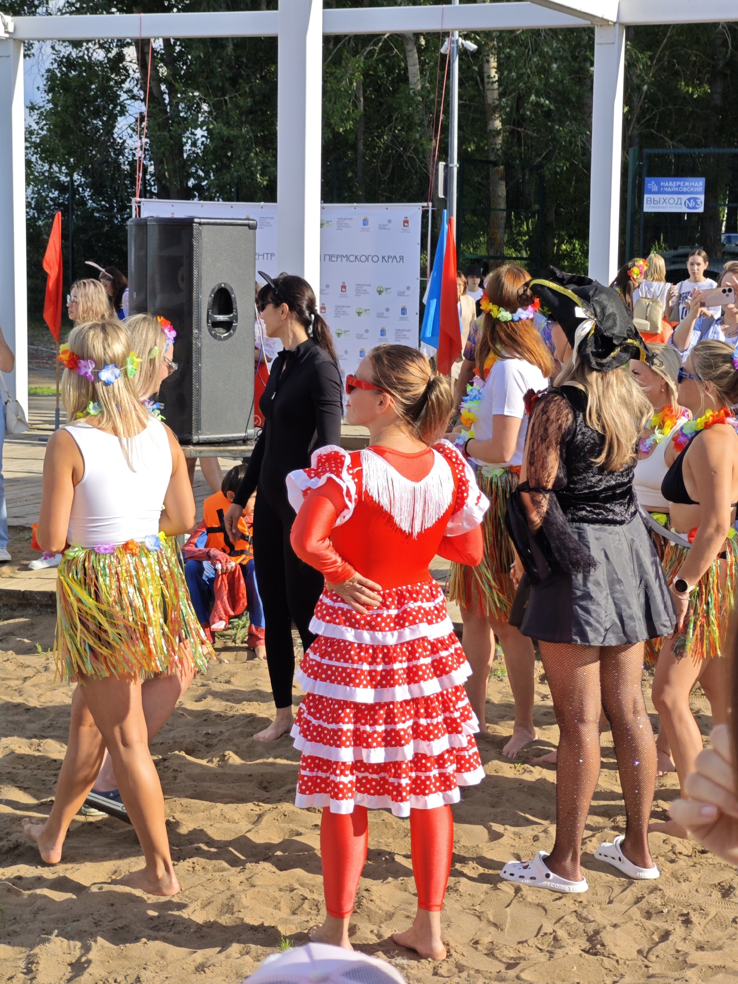 In this image, a group of people, mostly women, are gathered on a sandy area, likely a beach or park, in Chaikovsky, Russia. The subjects are dressed in festive, colorful attire, suggesting a celebratory event or a themed gathering. Several women are wearing leis and flower crowns, with some sporting hula skirts, indicating a possible tropical or summer theme. One woman is dressed in a red and white polka dot dress and red leggings, while another wears a black witch's hat and a black outfit with lace details. There's a large speaker on a stand, suggesting a DJ or a performance is part of the event.
In the background, there are trees and what appears to be a fence with signs. One sign, visible in English and Russian, reads "НАБЕРЕЖНАЯ ЧАЙКОВСКИЙ" (Embankment Chaikovsky) and "ВЫХОД №3" (Exit No. 3), confirming the location. Another sign in Russian has text that includes "ПЕРМСКОГО КРАЯ" (Perm Krai). The scene appears to be taking place during the daytime, under clear skies, with bright sunlight casting shadows on the sand. The mood seems lively and joyful, with people socializing and enjoying the occasion.