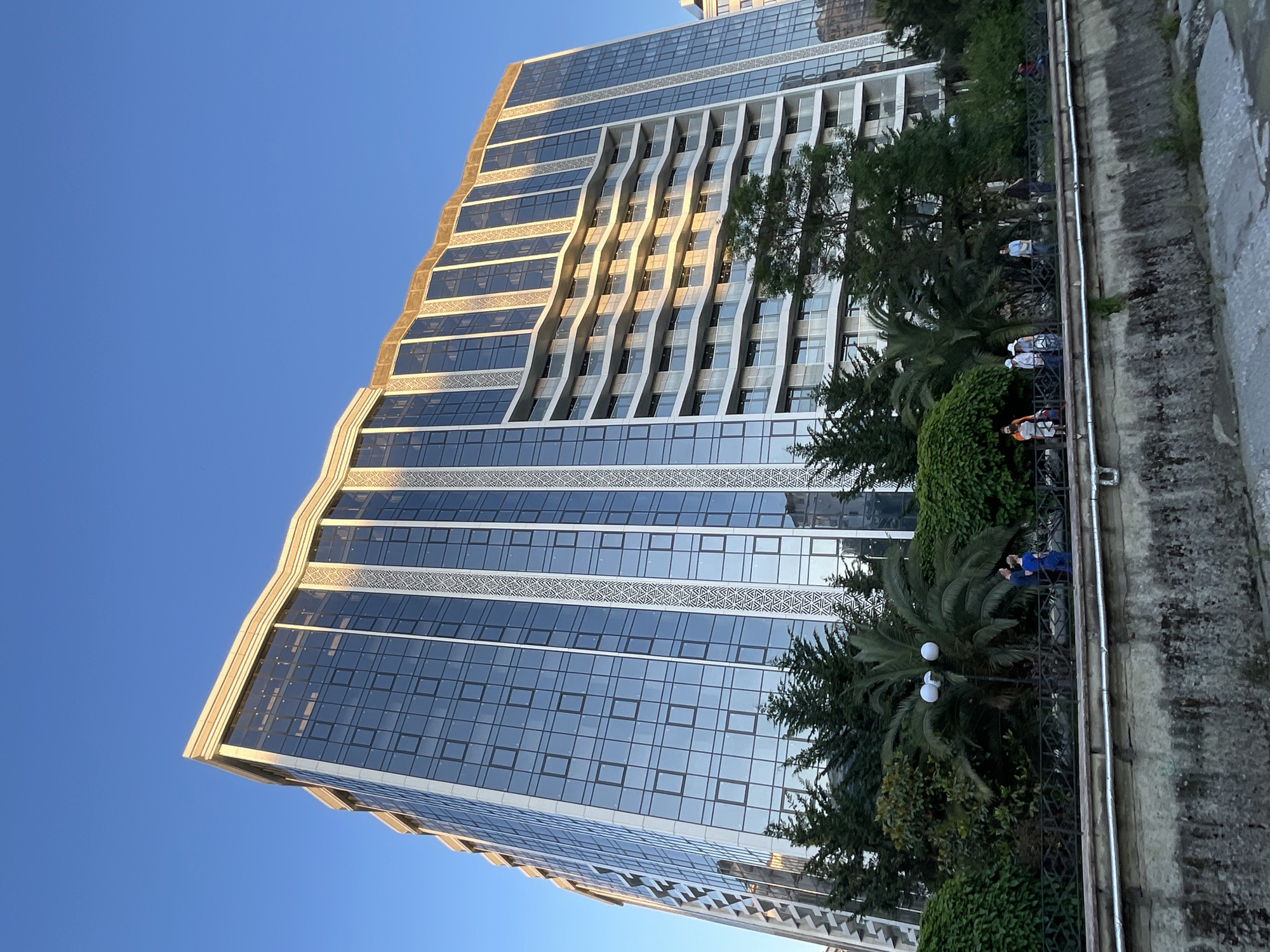 This image depicts the Apartment Hotel Imeretinsky, situated within the Imeretinsky Resort area of Sochi, Russia, under a clear blue sky. The prominent subject is a large, modern, multi-story building featuring a blue-tinted glass facade that reflects the sky. The building exhibits a distinctive architectural design with wavy, undulating elements along its roofline and on the numerous balconies that span several floors on one side. Vertical architectural bands with repeating geometric patterns are integrated into the facade.
In the foreground, a dense strip of lush green vegetation, including conifers, palm trees, and various shrubs, partially obscures the lower levels of the building. A dark, ornate metal fence runs horizontally across the midground, defining a public walkway. Along this walkway, approximately seven to ten individuals are visible, either standing or walking. They are dressed in casual attire; one couple is distinctively dressed in blue. Below the fence and walkway is a concrete or stone embankment featuring two horizontal metal pipes, suggesting the presence of a canal or riverbed just out of view at the bottom of the frame. Sunlight illuminates the upper portions of the building and the balconies, casting a golden hue on the light-colored structural elements.