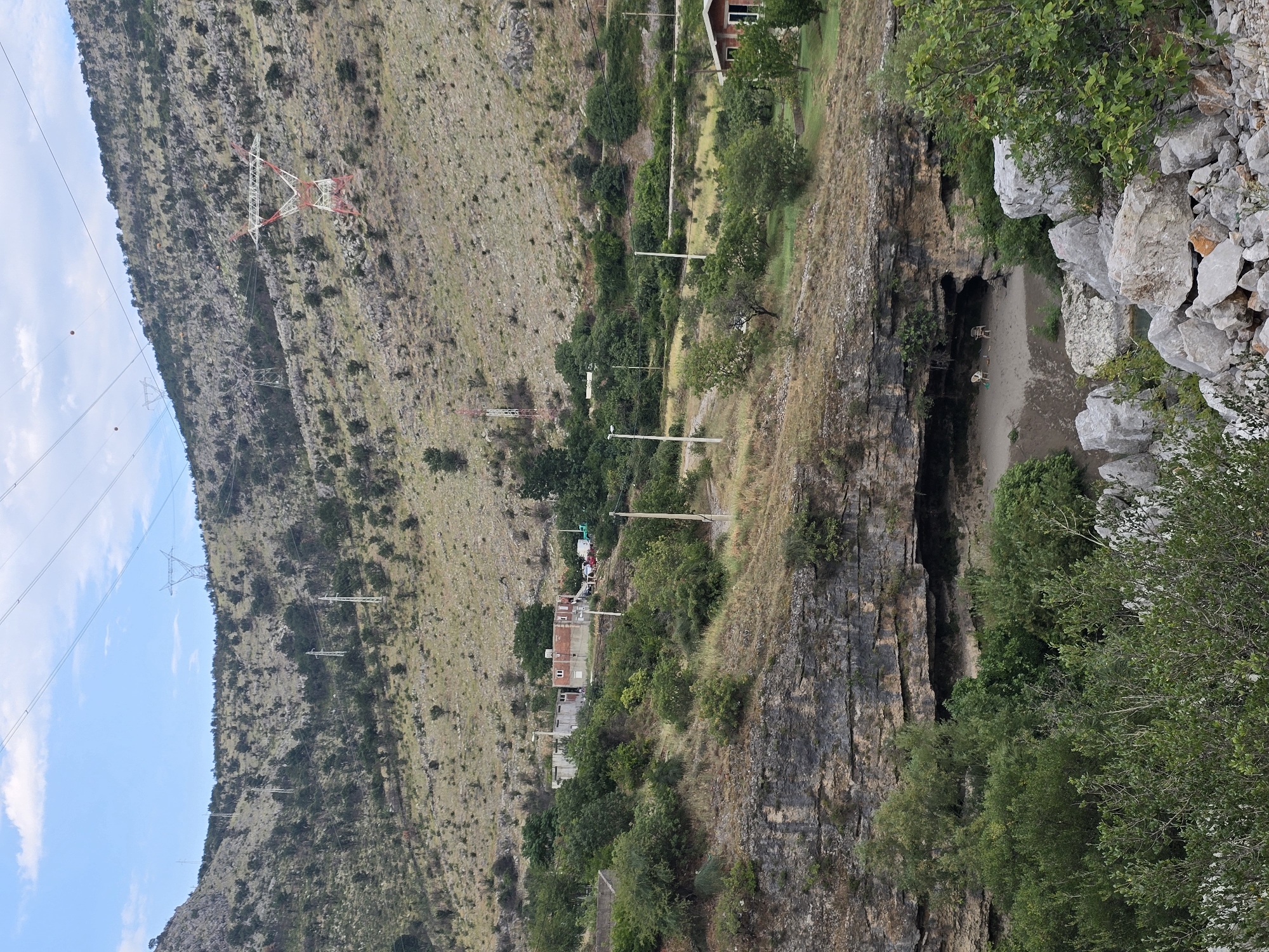 The image depicts a rugged, mountainous landscape in Podgorica, Montenegro. A prominent feature is a large cave opening in a rocky cliff face, revealing a sandy interior where two chairs are visible. The surrounding terrain is sparsely vegetated with scrub and trees, interspersed with rocky outcrops. Power lines and pylons traverse the hillsides, indicating human infrastructure in the natural environment. In the distance, a cluster of buildings, possibly residential or industrial, can be seen on the slope. The sky is partly cloudy, suggesting a temperate climate. The overall scene conveys a sense of remote, undeveloped wilderness coexisting with modern technological elements.