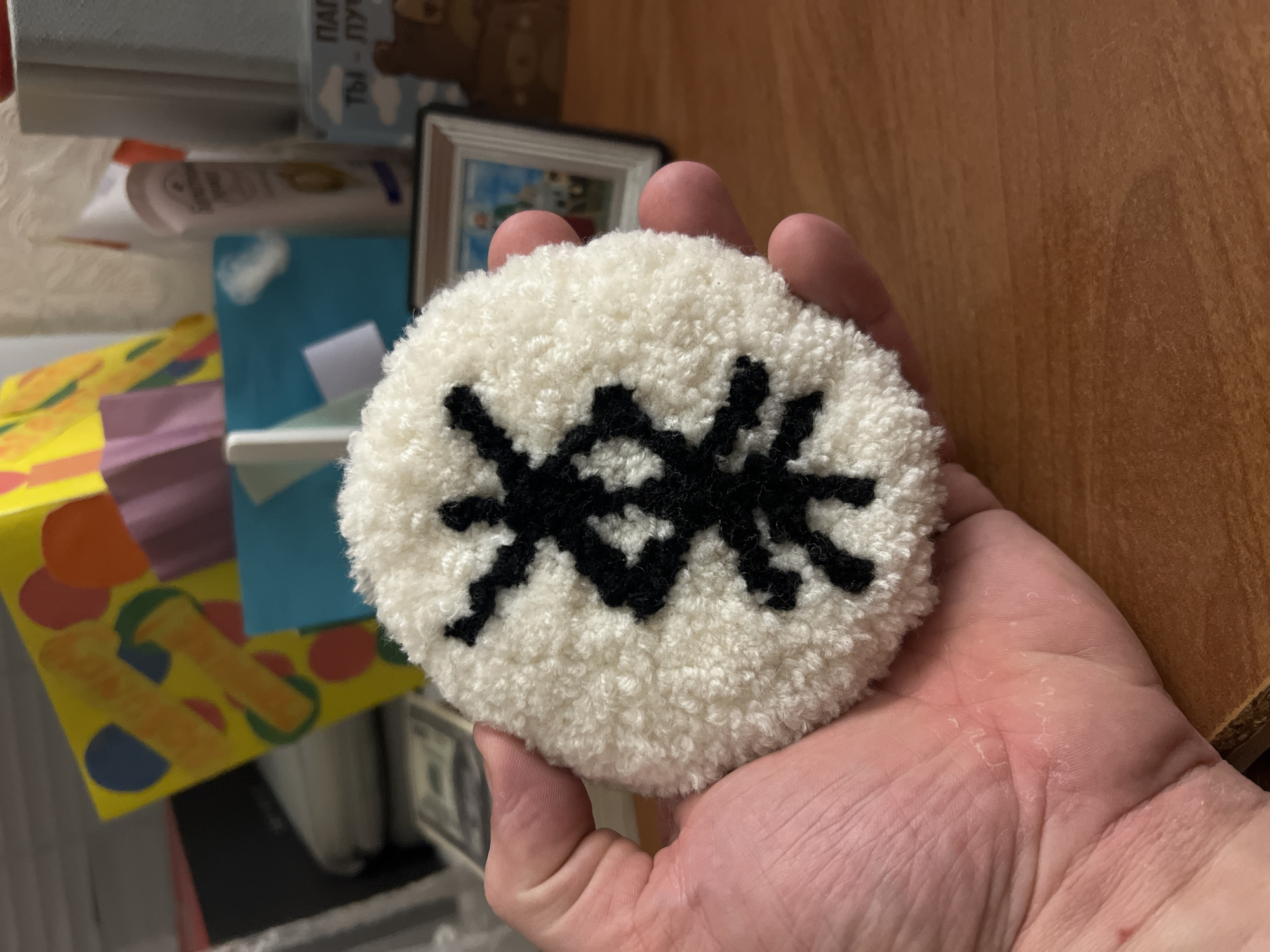 A close-up image shows a person's hand holding a circular, fluffy object with a black, spider-like design on its surface. The object appears to be a crafted item, possibly a coaster or decorative piece made from yarn or similar materials. The background is somewhat cluttered, showing parts of shelves or a desk with items like a picture frame, containers, and colorful papers or cards. The scene is likely indoors, but there is no specific information in the image to determine the exact location, particularly within the city of Yoshkar-Ola, Russia.