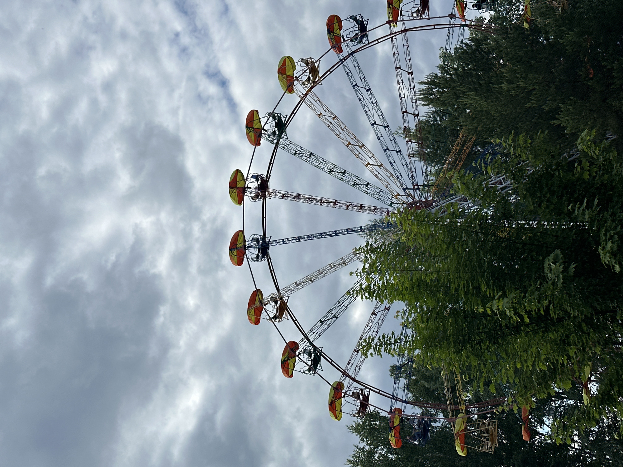 The image depicts a large Ferris wheel in an outdoor setting under a heavily overcast, grey sky in Unknown, Unknown. Dense green foliage from numerous trees occupies the foreground and midground, partially obscuring the lower half of the Ferris wheel's structure. The Ferris wheel is a metallic, multi-colored structure, with visible sections of red, yellow, and blue on its spokes and frame. It features multiple gondolas, each equipped with a distinctive, bicolored parasol-style roof, predominantly orange and yellow. Several gondolas are occupied by individuals, with at least five clearly showing one or more passengers seated. The people are indistinguishable beyond their presence as riders. The scene suggests an operational amusement park or fairground attraction.