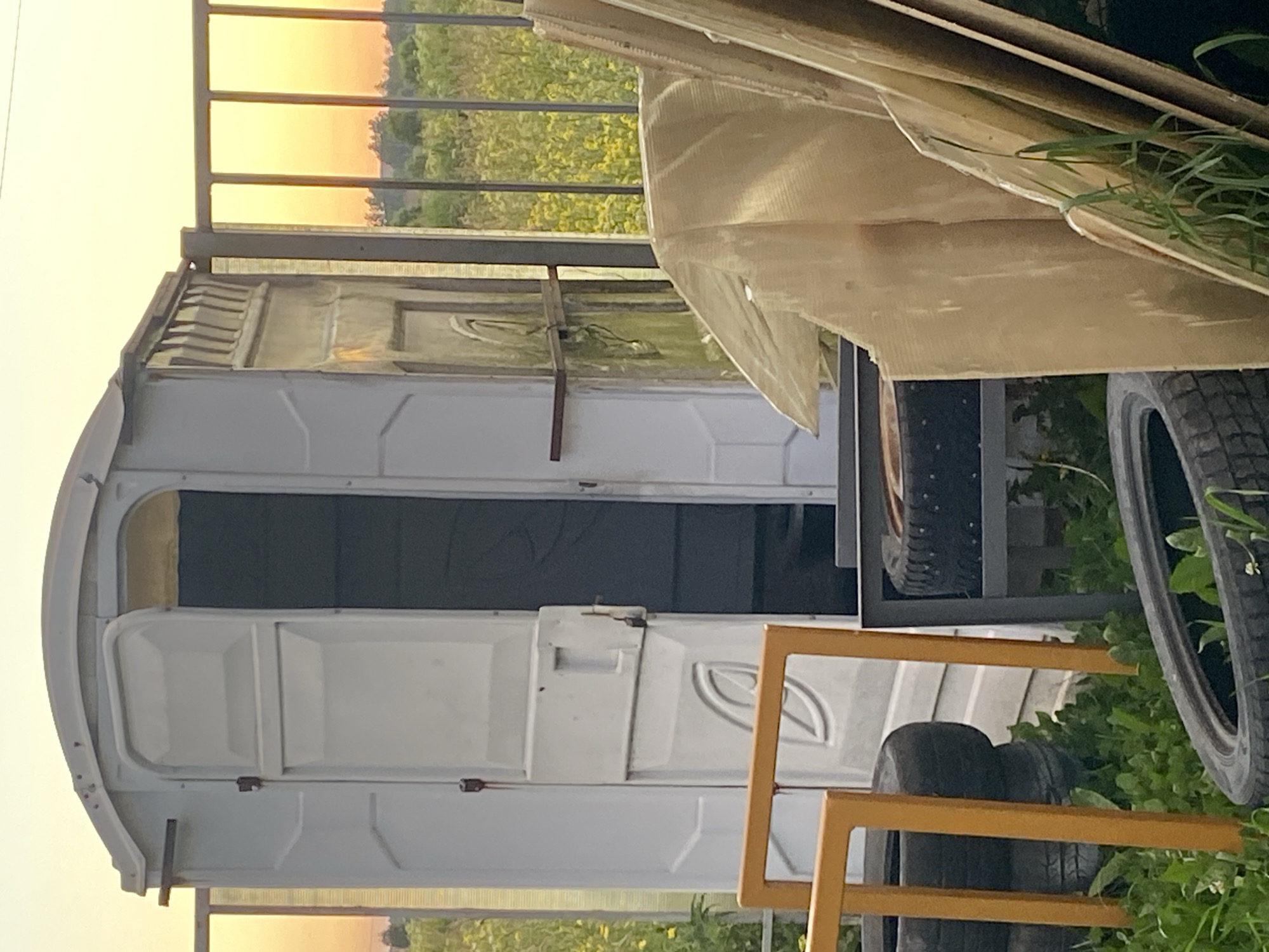 In Bol'shoe Zaborod'e, Russia, a portable toilet stands open next to a metal fence. The toilet is white with a gray interior. A metal shelf is attached to the side of the toilet. In front of the toilet are two black tires stacked on top of each other, supported by an orange metal frame. To the right of the toilet, several large flat pieces of wood are leaning against a dark metal frame, on which another tire rests. The scene is set against a backdrop of a field of yellow flowers and green vegetation under a pale orange sky. The sun is setting, casting a warm glow over the landscape.