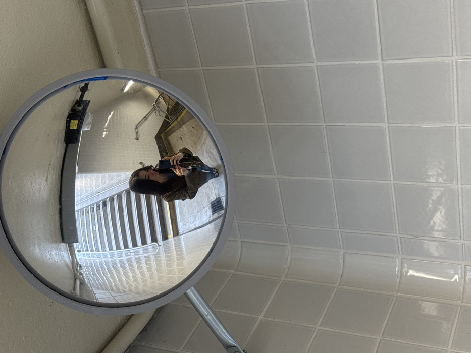 The image displays a large, circular convex mirror with a grey frame, mounted on a light-colored ceiling. The mirror reflects a single individual, appearing as a female with dark hair, wearing a dark olive-colored jacket over a lighter top, blue jeans, and light-colored footwear. A chain-link strap is visible over her right shoulder. She holds a dark-colored smartphone with both hands, positioned to capture her reflection in the mirror, with wired earbuds dangling from the device.
The background reflected in the mirror details a public stairwell within what appears to be a transit station. This area features a flight of light-colored concrete stairs with defined treads and risers, ascending towards the upper left from the reflected perspective. Metal handrails line the stairwell. The walls within the reflected area are clad in light-colored square tiles, matching the tiling in the immediate foreground. A small floor grate is visible near the base of the reflected stairs. Overhead in the reflection, a dark-colored piece of equipment or light fixture with a yellow label is mounted.
The immediate environment surrounding the mirror, from the perspective of the original photograph, consists of light grey or off-white square tiles with white grout, covering the walls and a curved corner. This specific architectural style, including the tiled passageways and stairwells, is characteristic of public transit infrastructure in the city. The location is consistent with a Port Authority Trans-Hudson (PATH) station in Hoboken, New Jersey, United States of America.