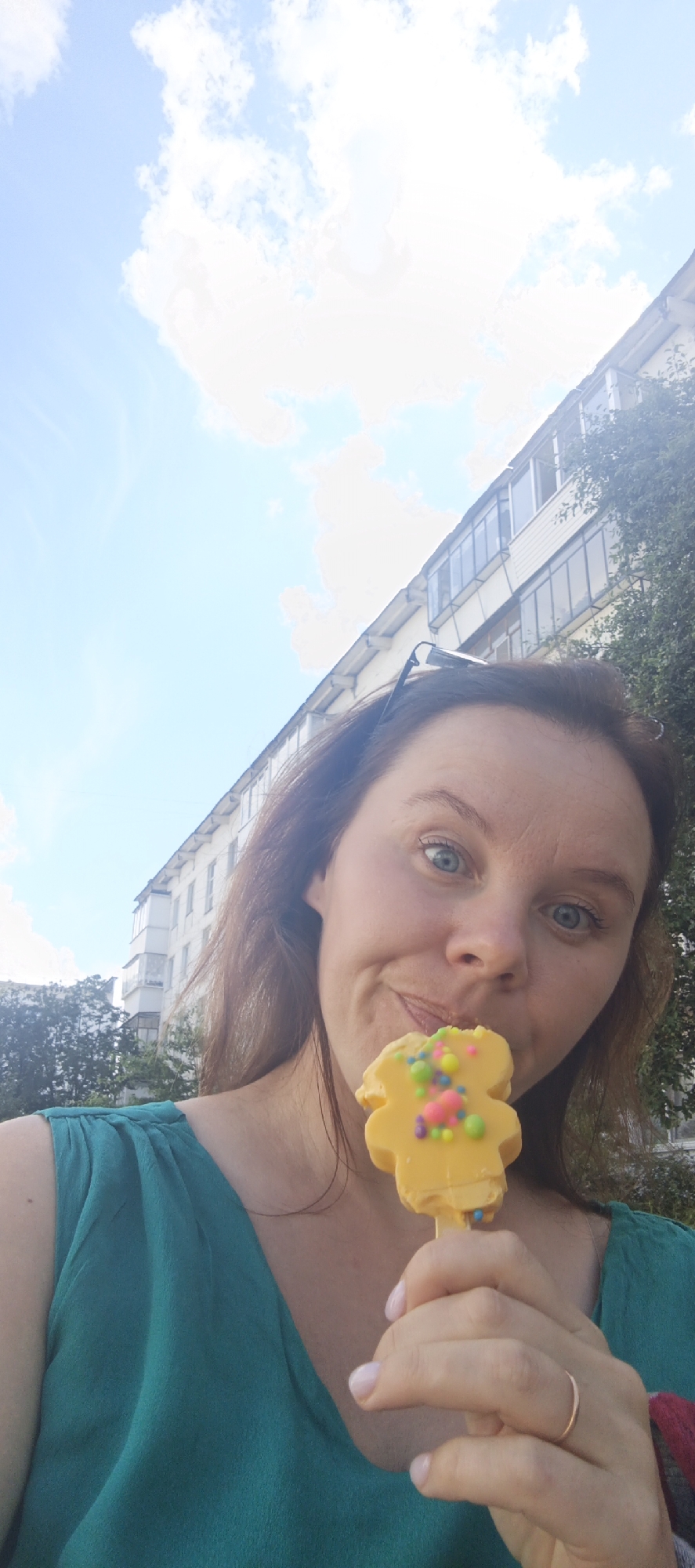 A woman, viewed from the chest up and from a low angle, holds a yellow, bear-shaped ice cream bar decorated with colorful sprinkles to her mouth. She is outdoors with a bright blue sky and white clouds above. Behind her, a multi-story apartment building with balconies and trees is visible. She is wearing a green, sleeveless top. She has blue eyes and brown hair. Her right hand, holding the ice cream, is raised. A thin gold ring is visible on her ring finger.