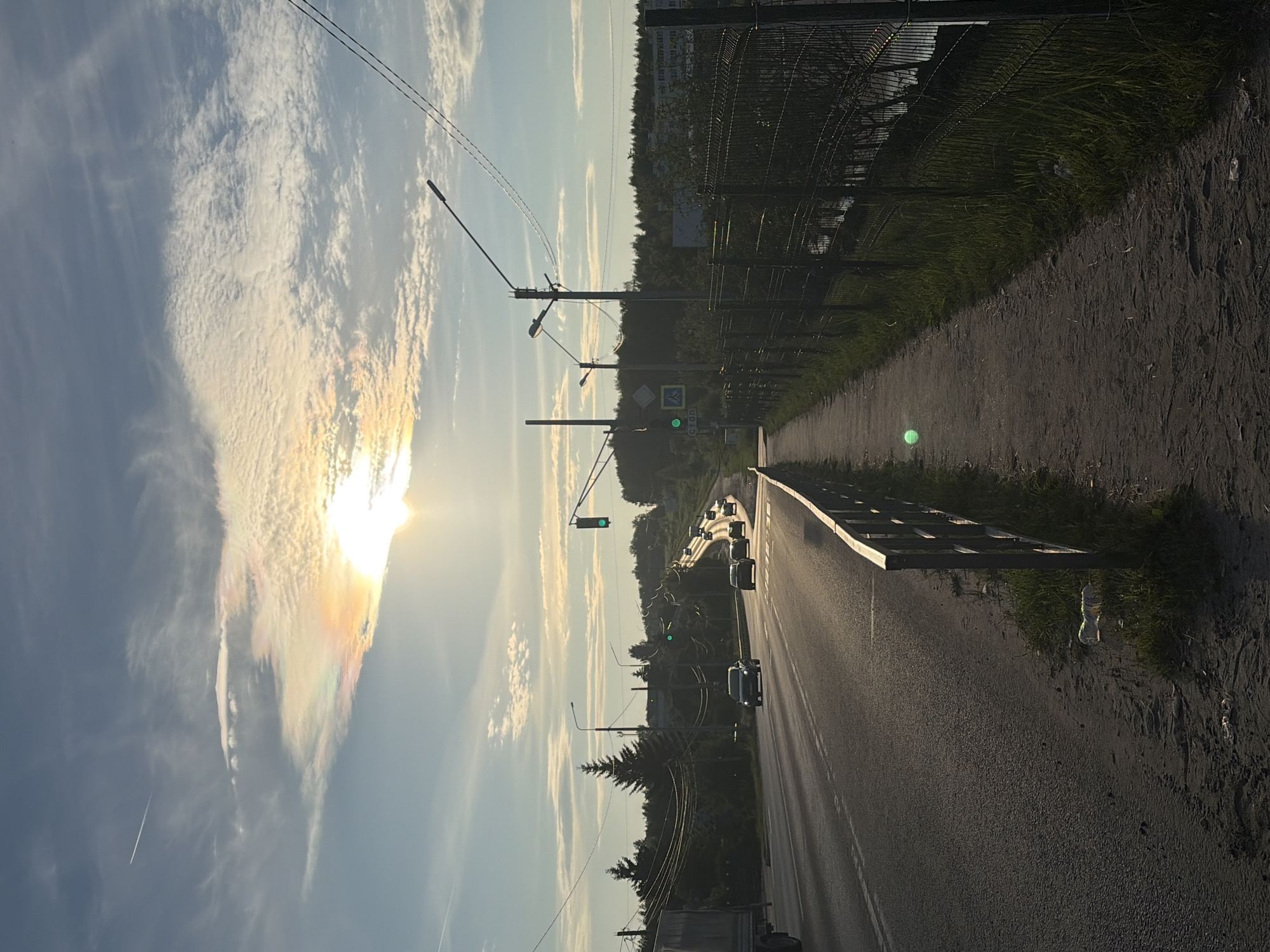 The image depicts a street scene in the village of Novinki, Russia, during daylight hours under a partly cloudy sky. The sun is low in the sky, casting a warm glow and creating a halo effect around it, possibly indicating a sundog or circumzenithal arc. A road stretches into the distance, curving to the right, with several cars traveling on it. A sidewalk runs along the right side of the road, bordered by a metal fence. The area is characterized by trees and vegetation, with some buildings visible in the background. Power lines and streetlights are present, indicating an urban or semi-urban environment. The ground on the sidewalk appears to be dirt and gravel, with some grass growing along the fence line. There is evidence of litter on the ground near the sidewalk. The overall impression is that of a roadside view in a rural or suburban setting at dusk or dawn.