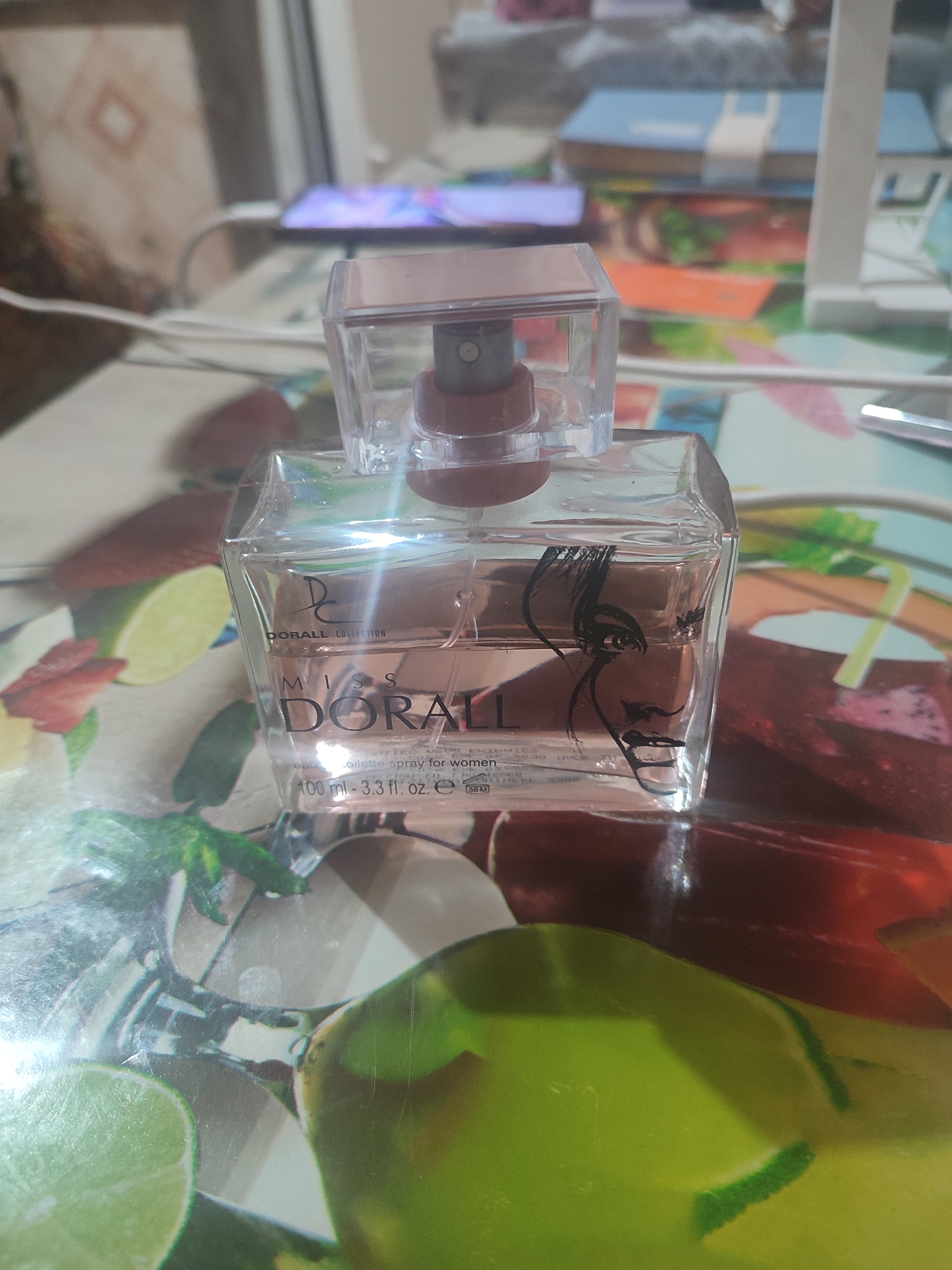 A close-up, eye-level shot shows a partially filled bottle of "MISS DORALL" eau de toilette spray for women, with a capacity of 100 ml (3.3 fl. oz.). The bottle is made of clear glass and has a square cap. The label on the bottle features the brand name "DORALL COLLECTION" above "MISS DORALL." A black, stylized drawing of a woman's face is visible on the side of the bottle. The bottle is positioned on a surface with a colorful, abstract pattern that includes images of fruit slices, such as limes and strawberries. In the background, a blurred image of a light-colored shelf with books and a smartphone displaying a colorful screen can be observed. The lighting suggests an indoor setting. No specific location in Chernivtsi, Ukraine, can be determined from the image.