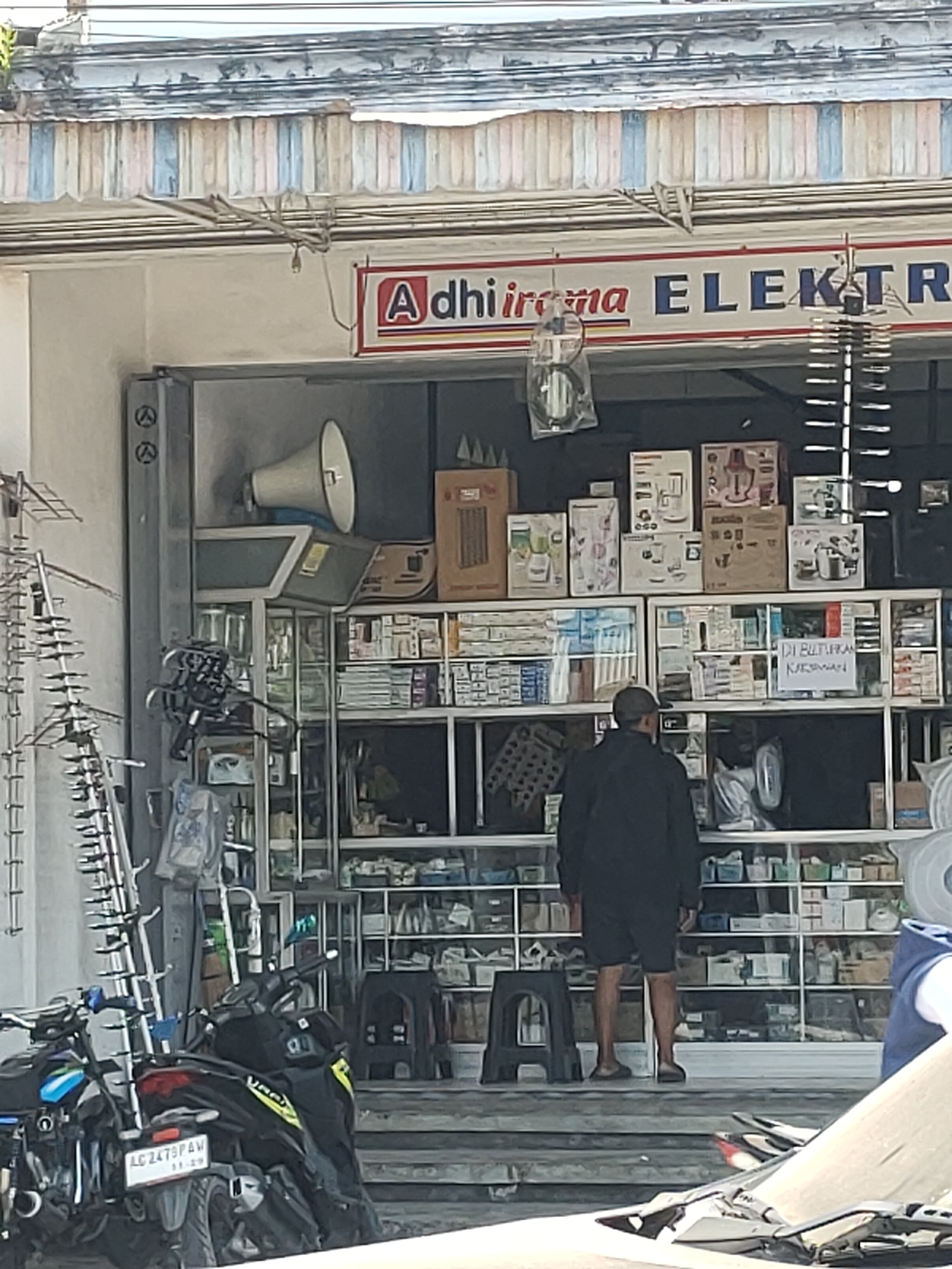 In Kanigoro, Indonesia, an outdoor view captures the storefront of an electronics shop named "Adhirama Elektra." The shop features a prominent sign with its name in red and blue lettering above a display of various electronic appliances and parts. Inside, shelves are stocked with boxed items, likely appliances, and smaller packaged goods. A man wearing a dark jacket and shorts stands facing the shop, his back to the camera. To the left of the entrance, a large megaphone is visible. In front of the shop, several motorcycles are parked, suggesting pedestrian and vehicular access to the establishment. The overall scene depicts a commercial area with typical street activity.