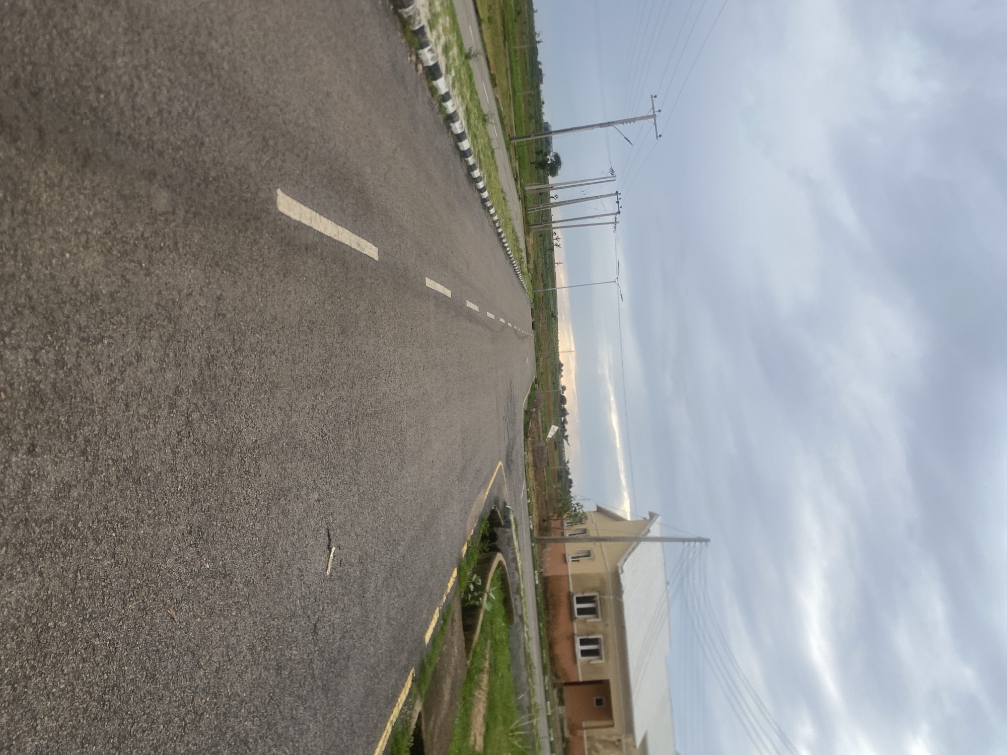 The media file depicts a street scene in Bulamare, Nigeria. The foreground is dominated by an asphalt road with white dashed lines marking lanes. To the left of the road, there is a curb with alternating black and white markings, followed by a strip of grass and then more of the road. In the distance, on the right side of the road, a modern residential building with beige and orange-colored walls and multiple windows is visible. Beyond the building, there are utility poles with overhead electrical wires stretching across the landscape. The sky is overcast with a mixture of gray and white clouds, suggesting a cloudy or possibly hazy day. The time of day appears to be late afternoon or early evening, given the subdued lighting. The scene is quiet and appears to be in a developing residential area, with the road suggesting accessibility and infrastructure. There is no visible text in the image.