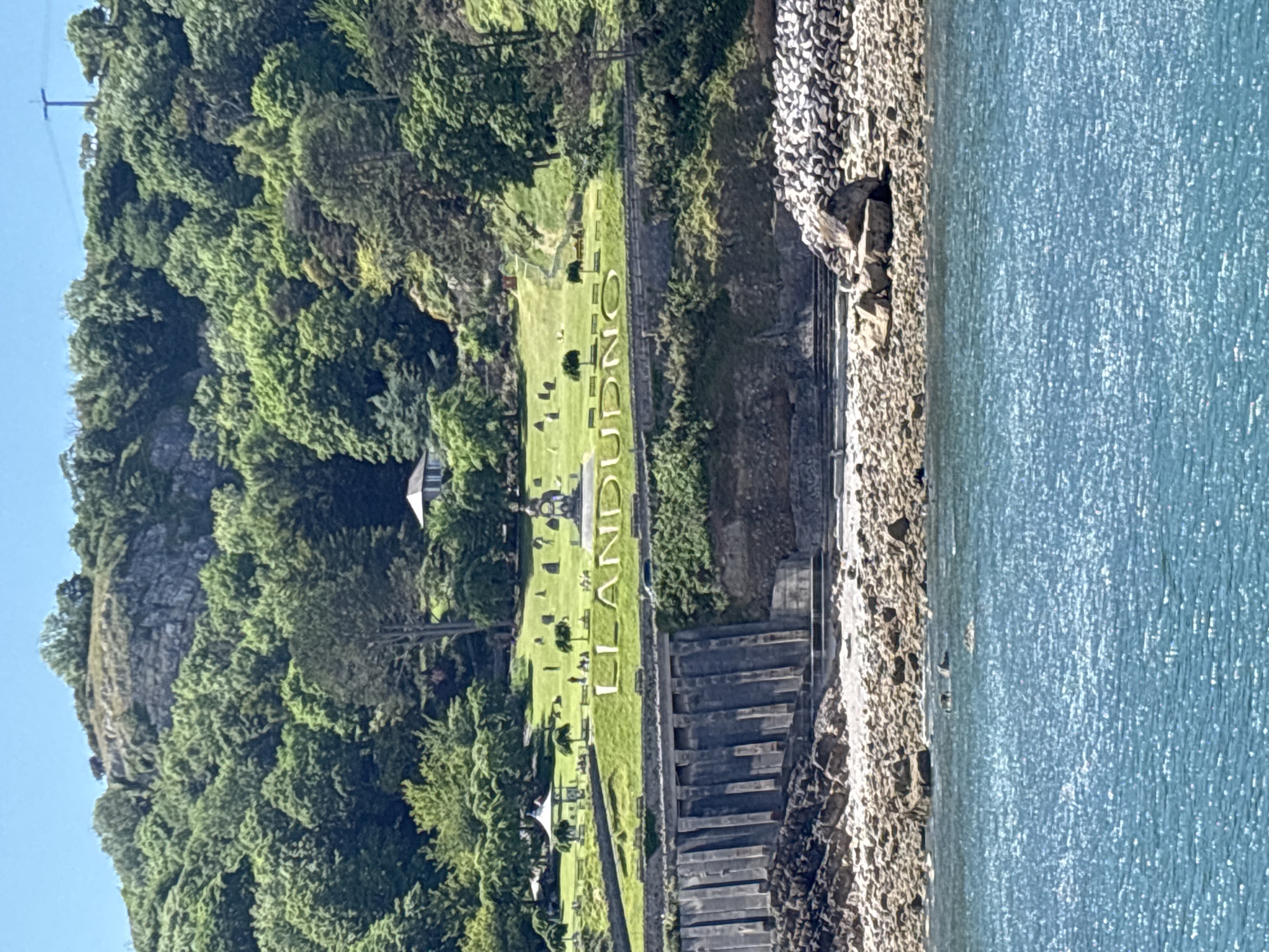 This is a landscape photograph taken in Llandudno, United Kingdom, featuring a hillside prominently displaying the town's name in large white letters on a grassy slope. The hillside is covered with dense green trees and vegetation. At the base of the slope is a stone wall with a staircase leading down to the water's edge. The water is a light blue color and appears to be part of the sea or ocean. The photograph is taken from an elevated position, looking down at the scene.