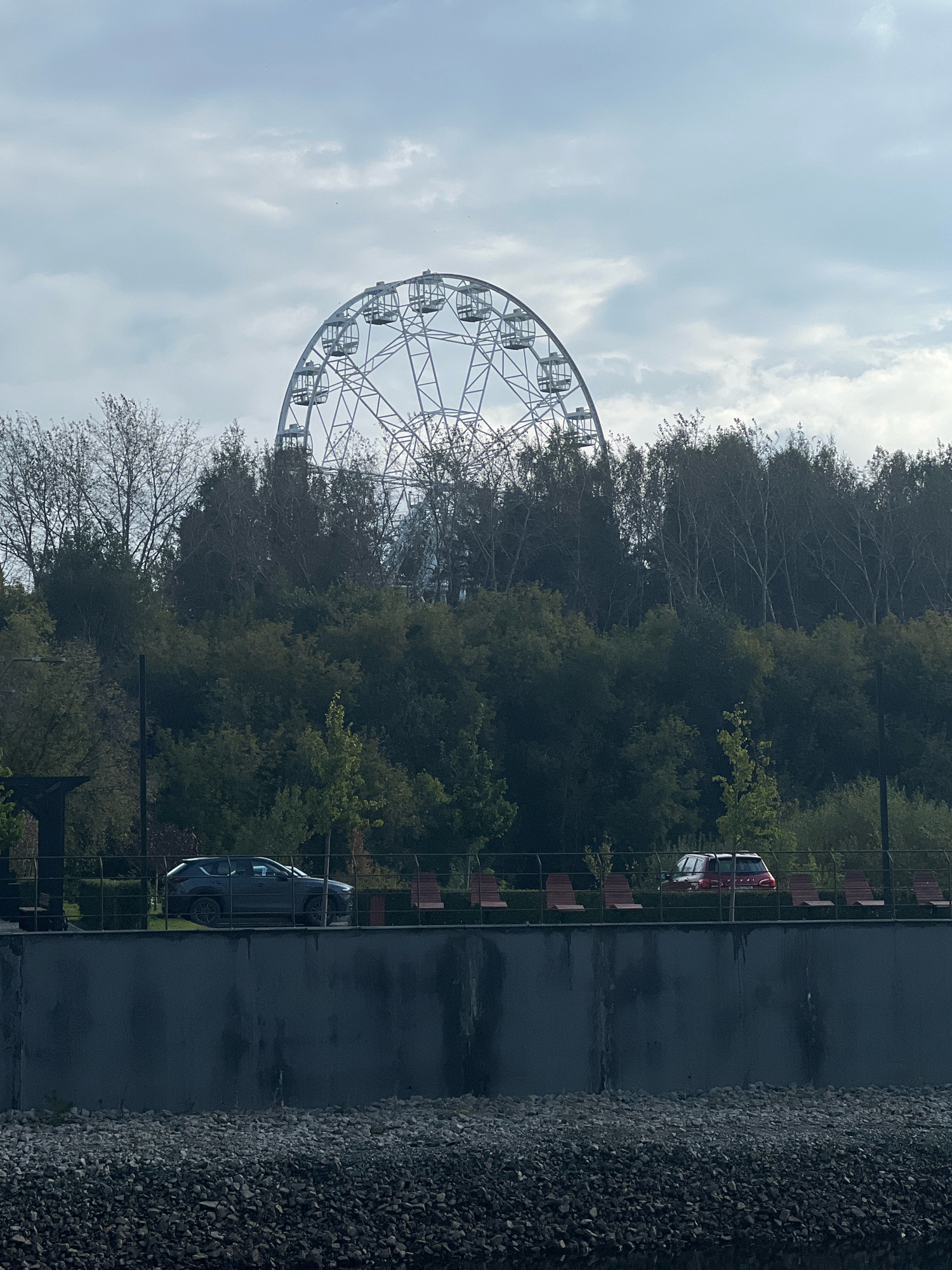 The image depicts a Ferris wheel towering over a dense line of trees. In the foreground, a dark gray car is parked on a paved area behind a metal fence. Several empty wooden benches are visible along the same paved area, also behind the fence. A dark, textured wall runs along the bottom of the frame, and a rocky embankment is visible below that. The sky is overcast with a pale blue hue. This scene is located in the city of Yekaterinburg, Russia, at the "Ural Mountains" Amusement Park, specifically near the Ferris wheel known as "Heart of Yekaterinburg".