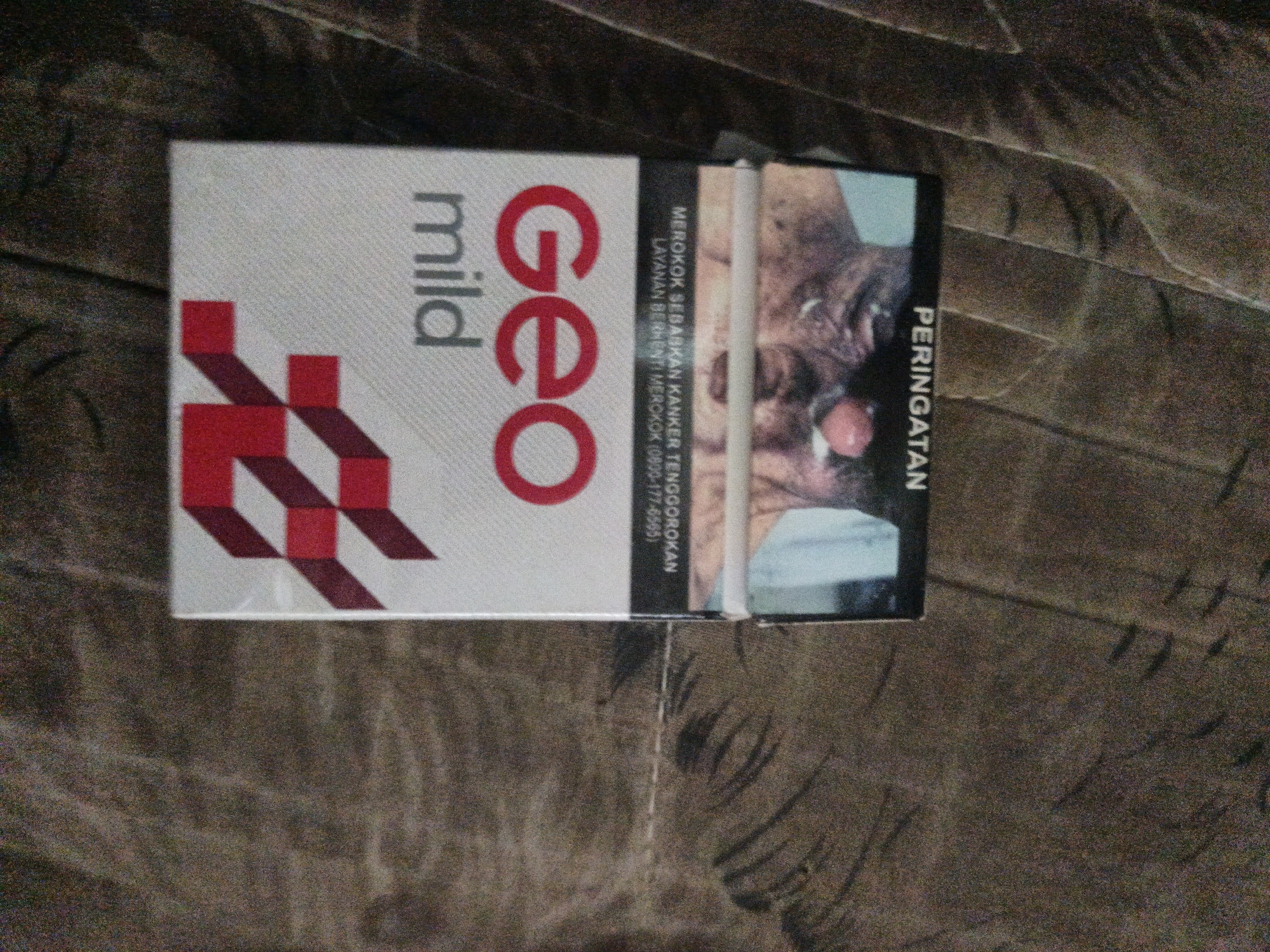 A pack of Geo Mild cigarettes is placed on a wooden surface, likely a table. The pack is mostly white with red and maroon accents. The front of the pack displays the brand name "GEO" in large red lettering and "mild" in smaller grey lettering. A red and maroon geometric design is also visible on the front. The side of the pack features text in Indonesian, including a warning about smoking causing cancer. The image of a person's face with a graphic element is present on the right side of the pack, which is a common feature on cigarette packaging for health warnings. The wooden surface appears to be a table with visible wood grain and texture. The lighting suggests an indoor environment. The location is identified as Temuwulan, Indonesia.