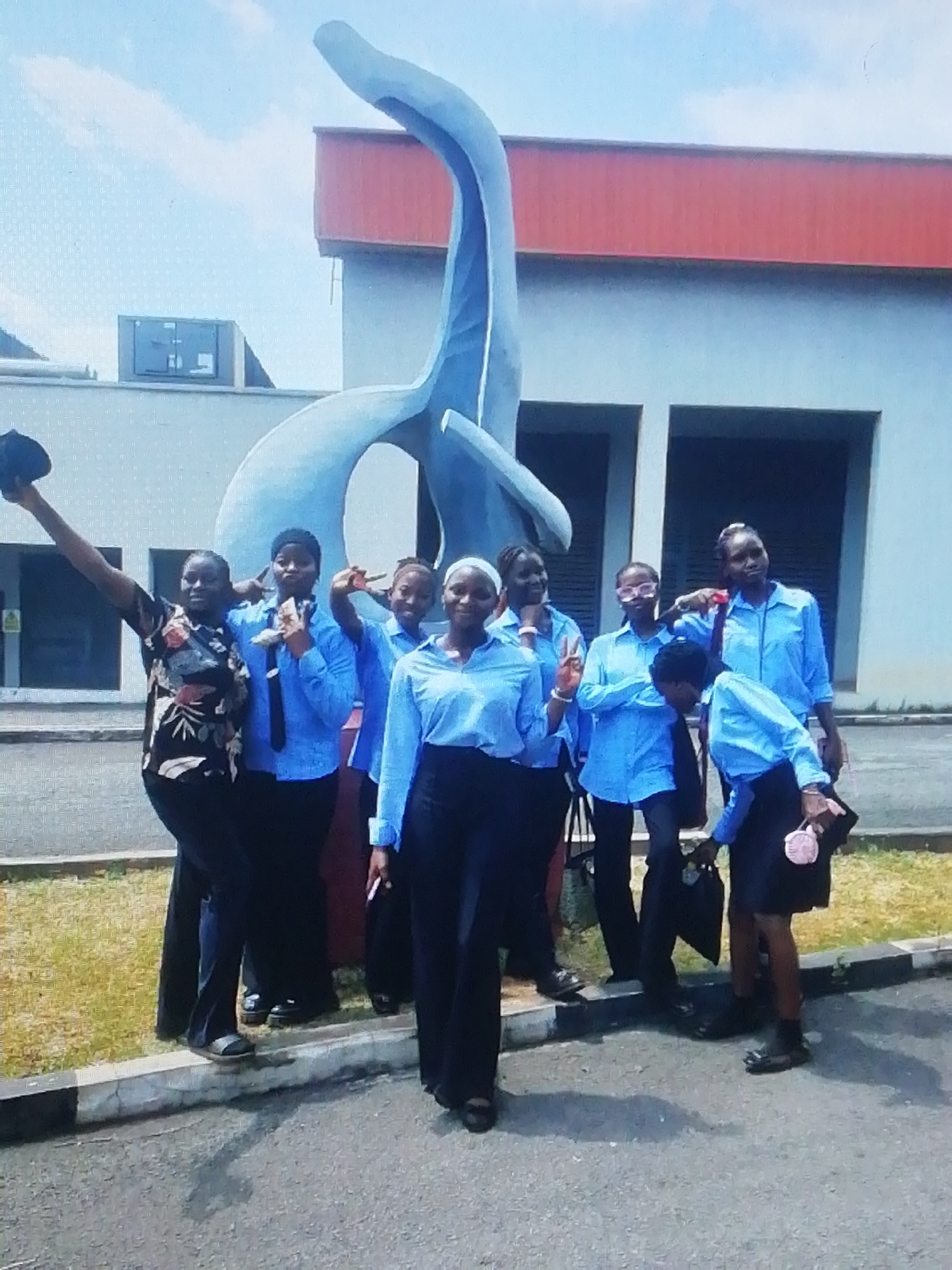 A group of nine people stands in front of a large, abstract, light-blue sculpture outside a light-colored building with an orange roof. The building has a row of rectangular windows. The people, mostly young women, are dressed in what appears to be school uniforms: light-blue shirts and dark pants or skirts. One woman wears a floral-patterned shirt and dark pants and holds a black hat in her raised right hand. Various individuals make hand gestures, including a peace sign. The background includes a clear sky. The scene is a gathering of people taking a photo in front of the sculpture. The location is the University of Nigeria, Enugu Campus.